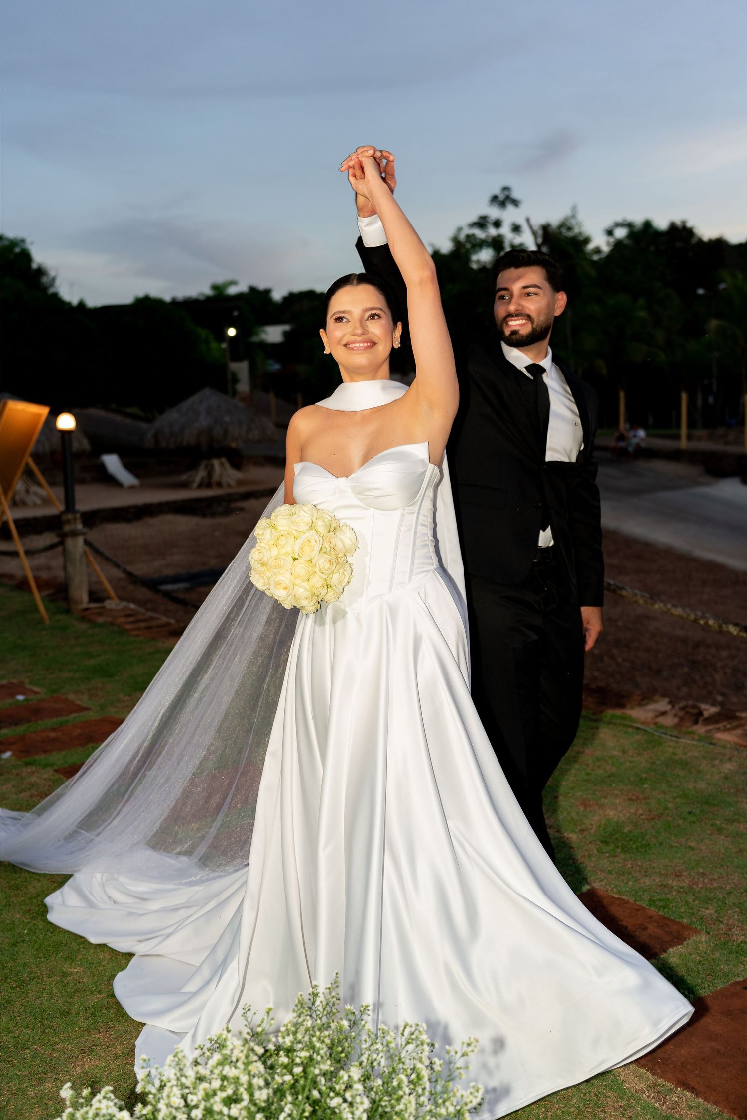 Bride and groom celebrate outdoors at dusk, holding hands raised, bride in white satin gown holding yellow bouquet.