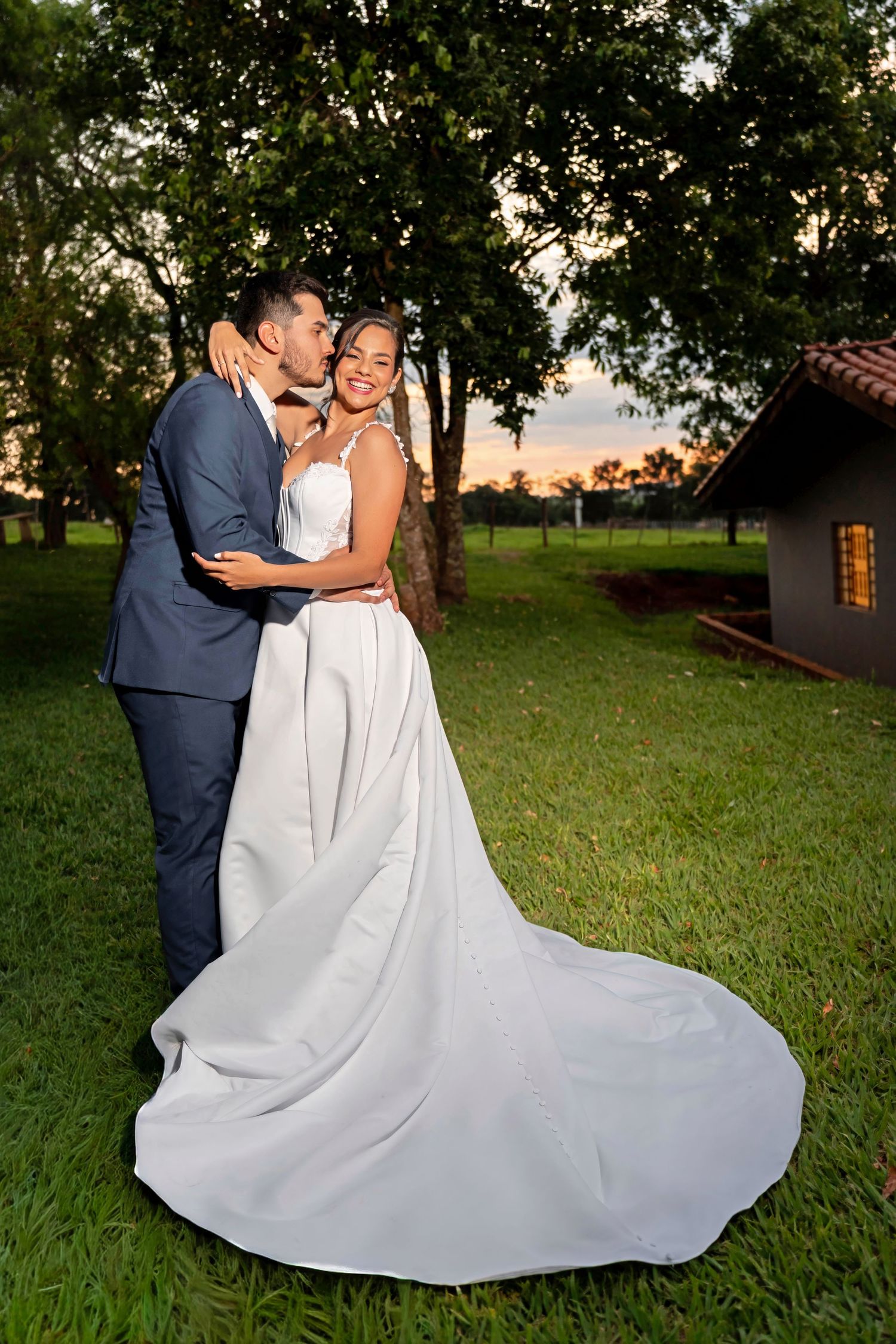 Bride in white gown with train and groom in navy suit share a romantic kiss outdoors at sunset.