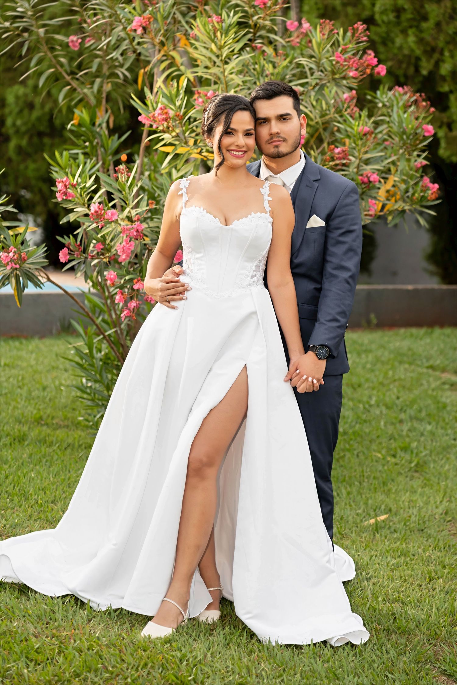 Bride in white slit gown and groom in navy suit pose together outdoors near pink flowering bushes.
