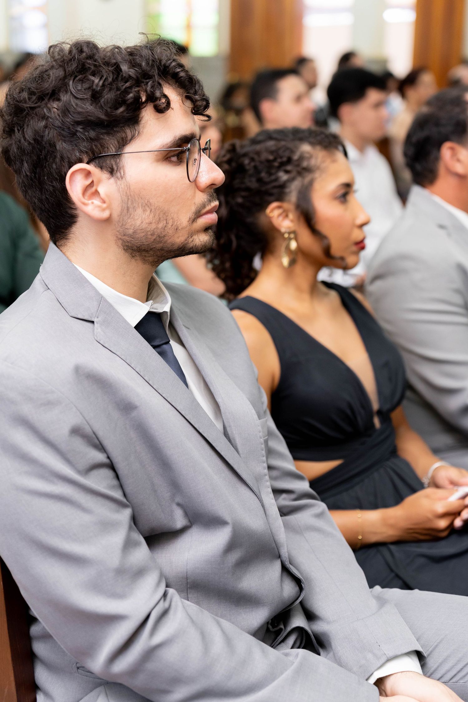 Guests seated in church pews during a wedding ceremony, man in gray suit and glasses in foreground.
