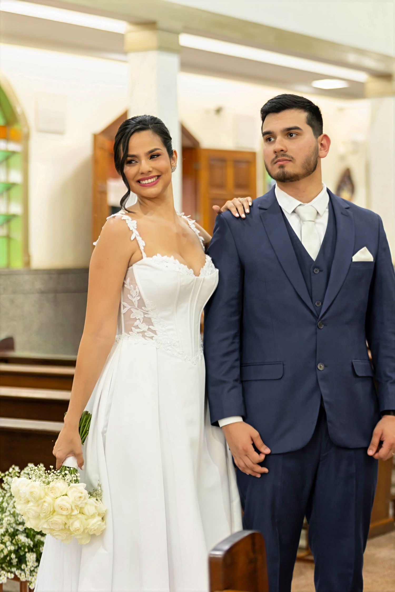 Bride in white lace gown holding bouquet stands with groom in navy suit inside a church.