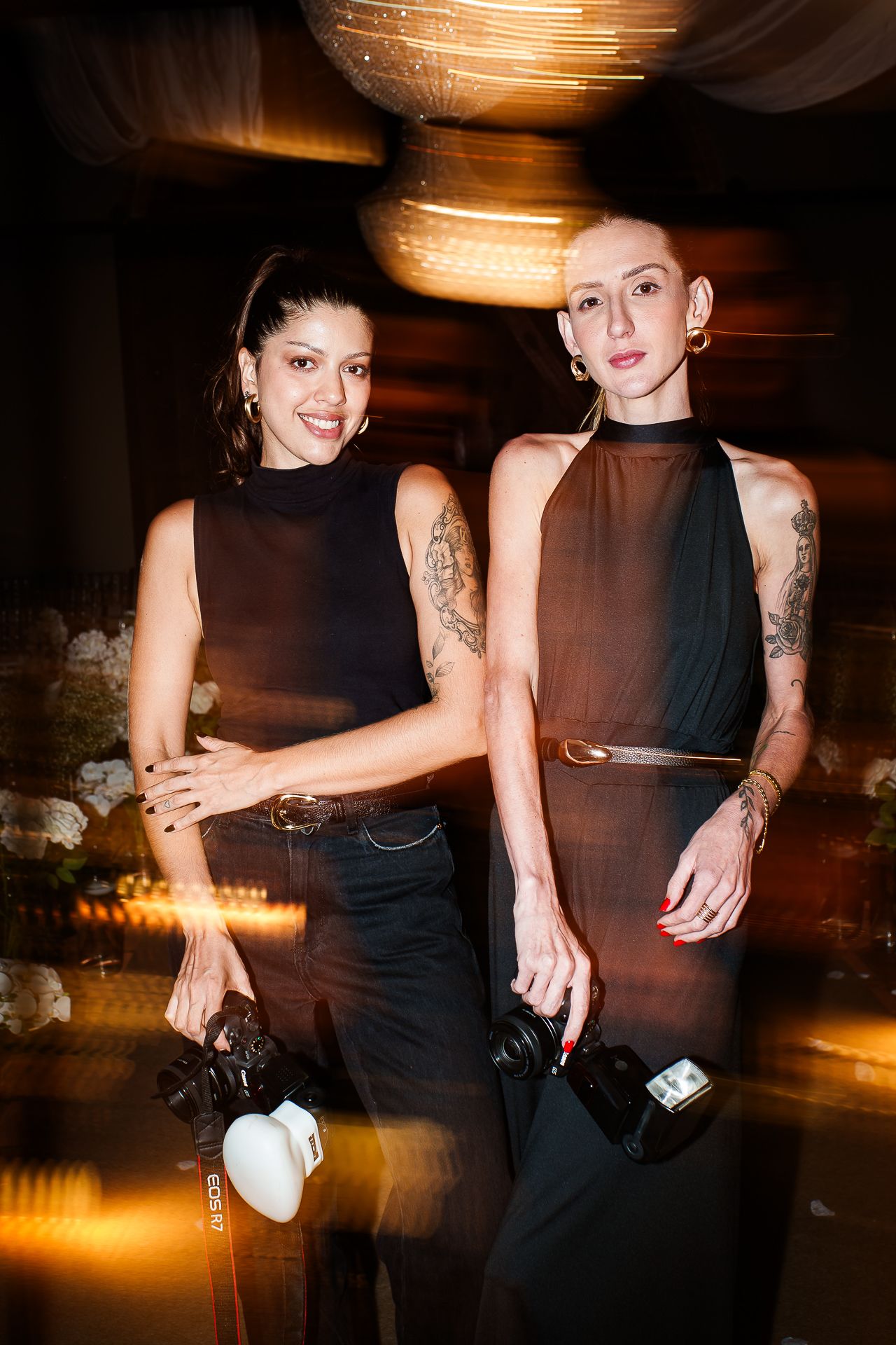 Two tattooed women in black outfits holding cameras pose together at an elegant evening event.