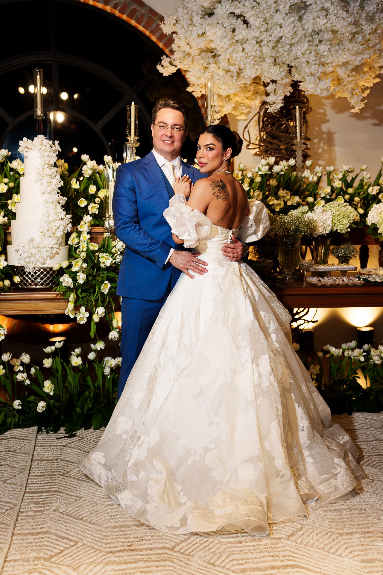 Bride in white ballgown and groom in blue suit pose together at elegant wedding reception with floral decor and tiered cake.