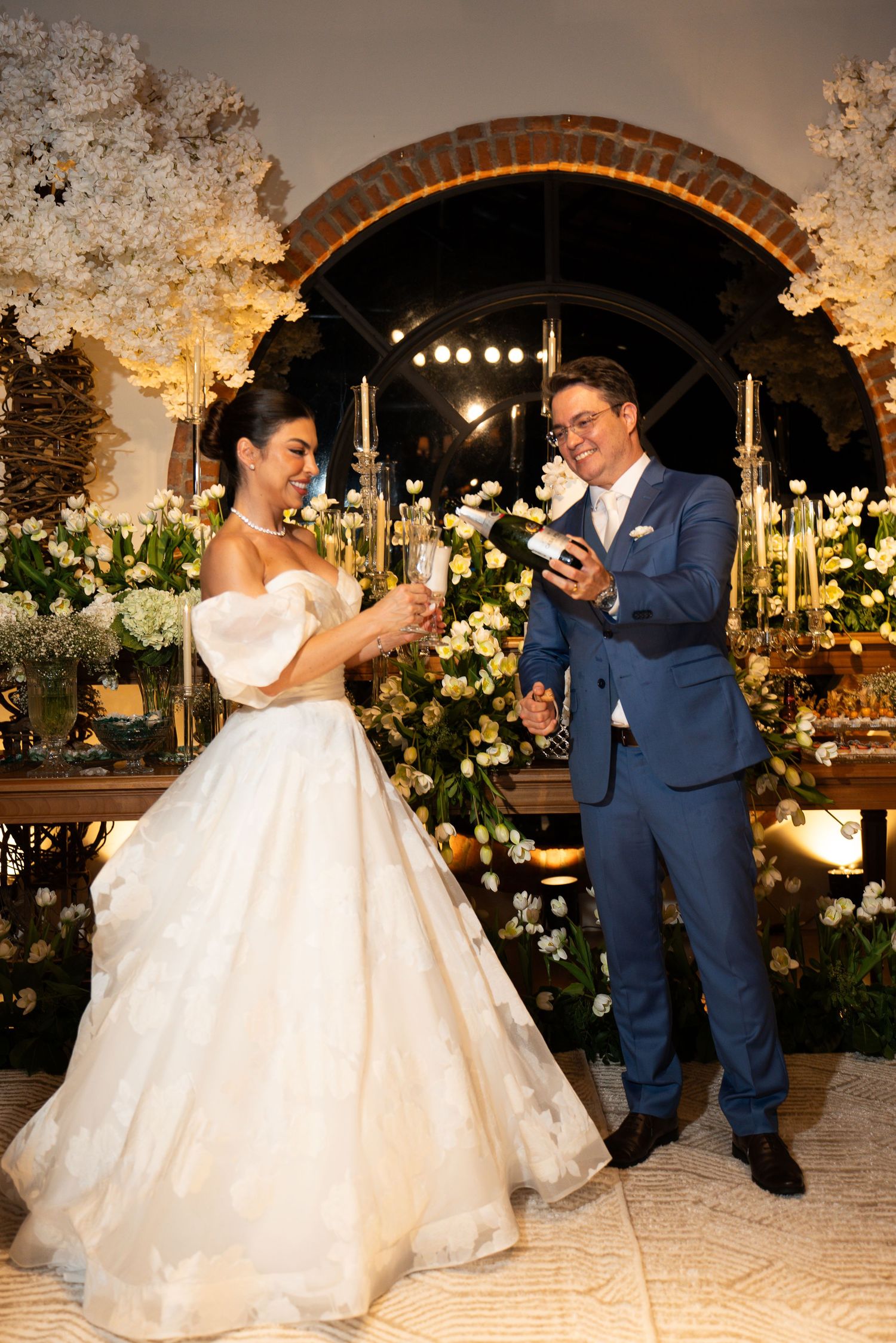 Bride in white ball gown and groom in blue suit toasting with champagne at elegant floral wedding reception.