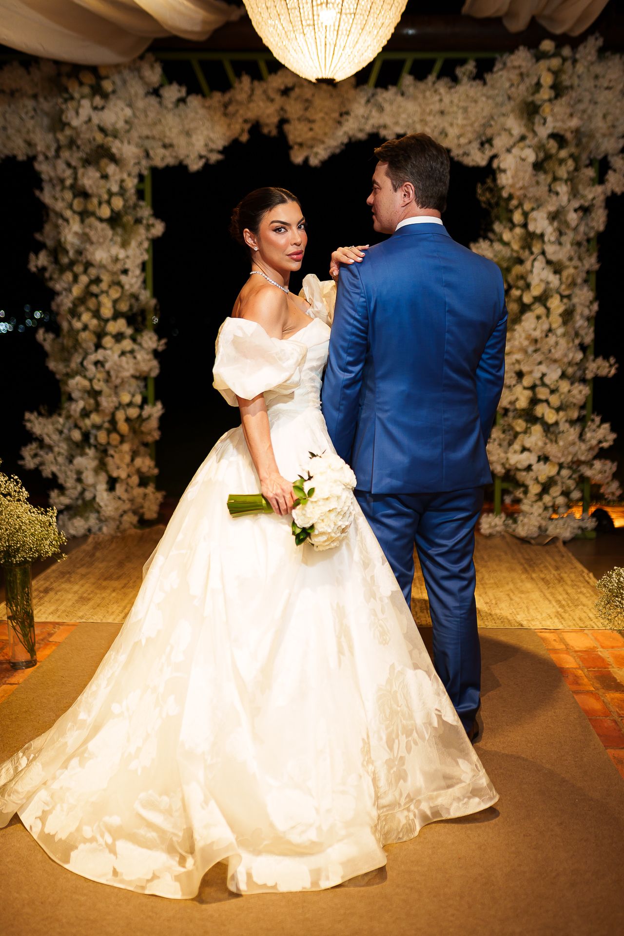 Bride in white off-shoulder ballgown holds bouquet beside groom in blue suit before floral arch at elegant wedding ceremony.