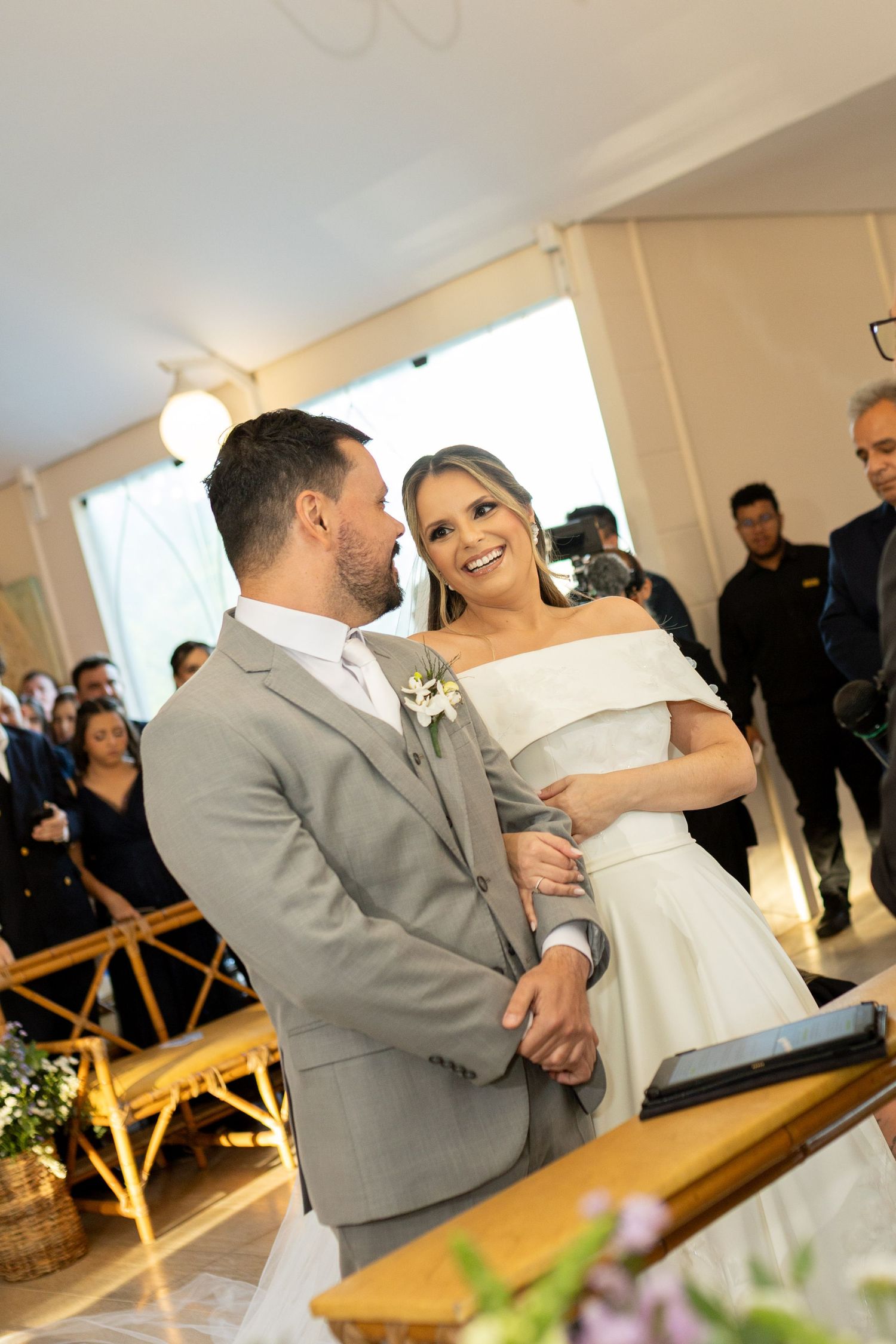 A smiling bride in an off-shoulder white dress and groom in gray suit exchange vows at their wedding ceremony.