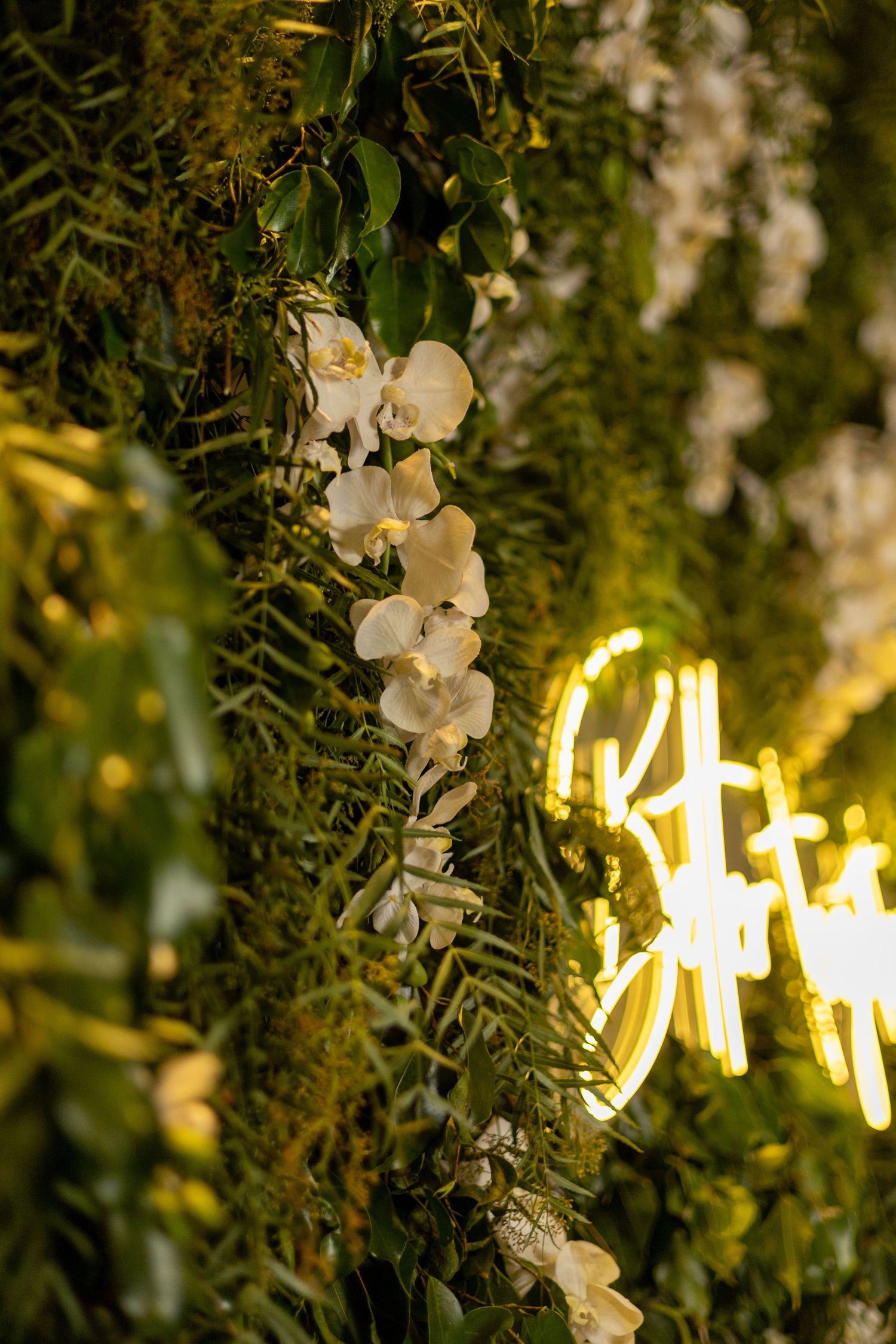 White orchids and lush greenery adorn a floral wall with glowing yellow neon sign at an elegant event.