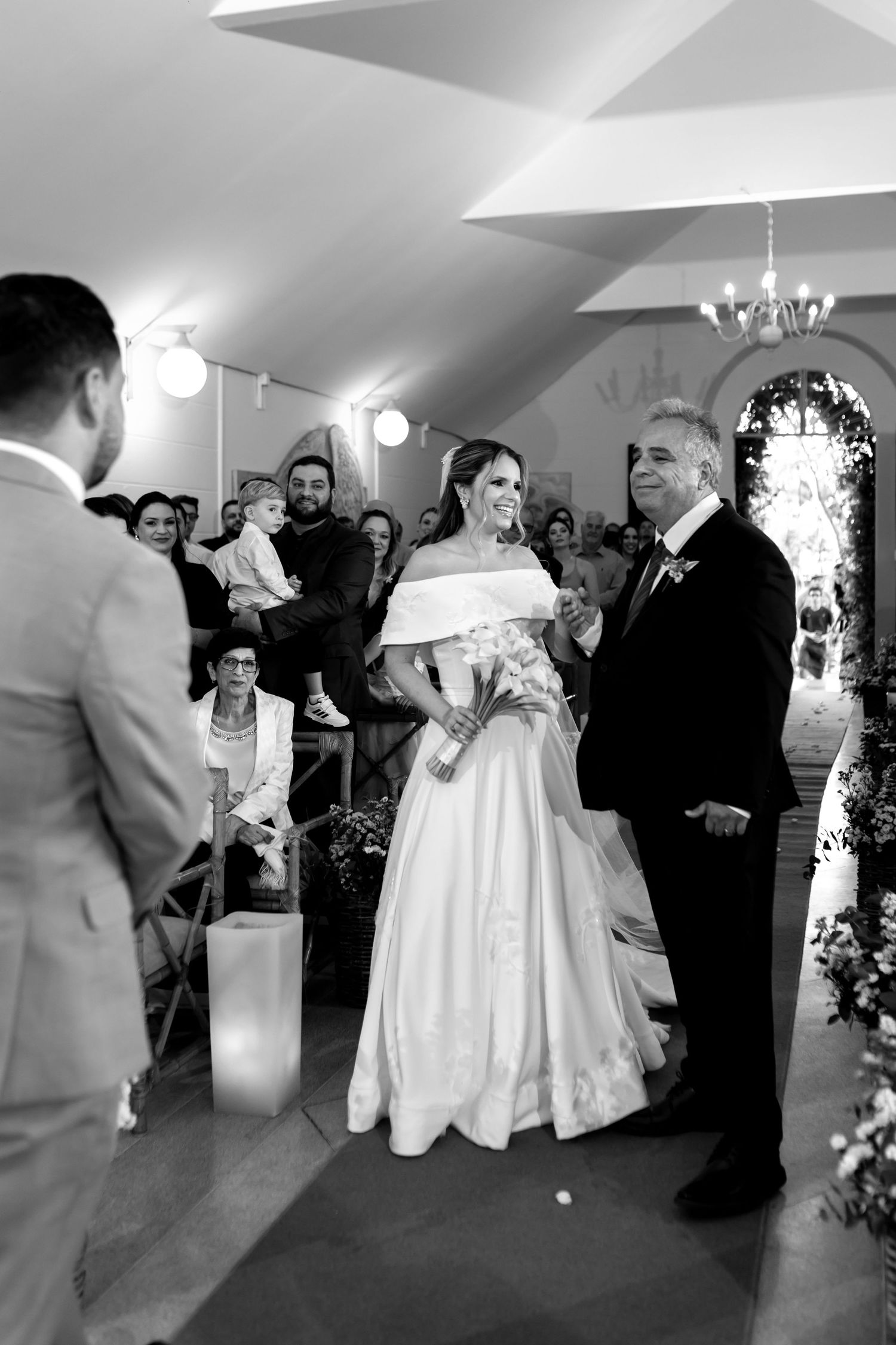 Black and white wedding photo of bride in off-shoulder gown walking down aisle with escort toward groom.