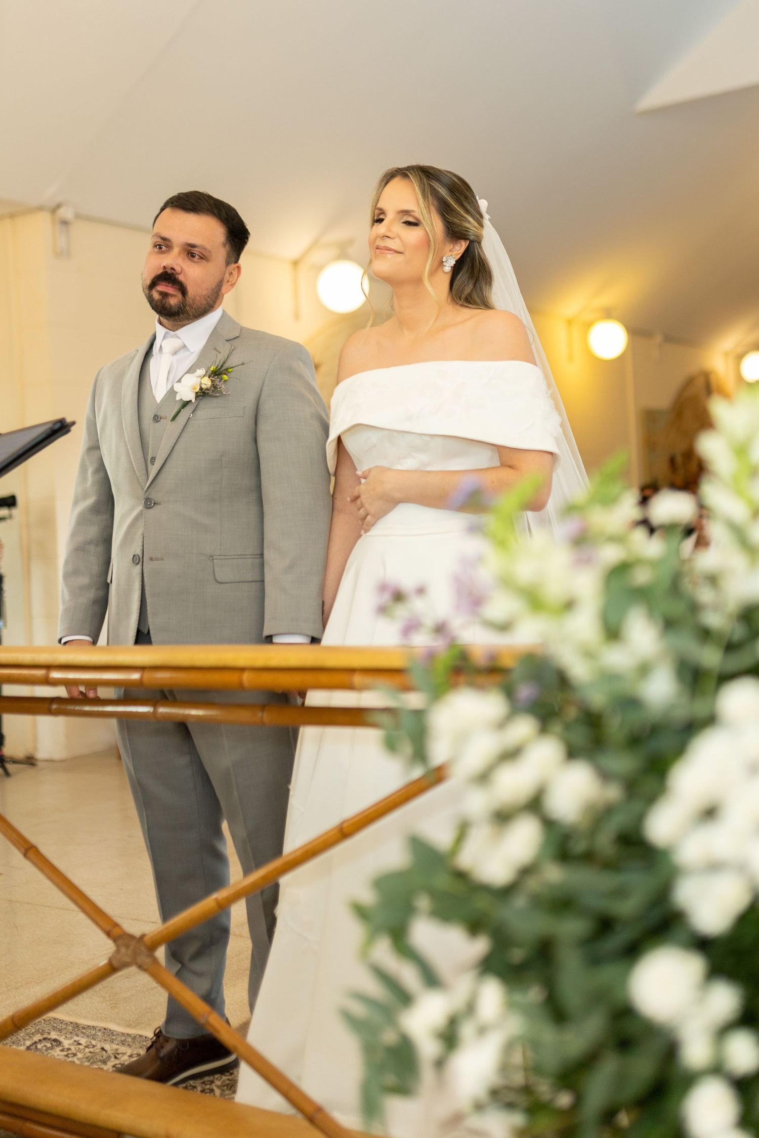 Bride in white off-shoulder gown and groom in gray suit stand at the altar during their wedding ceremony.