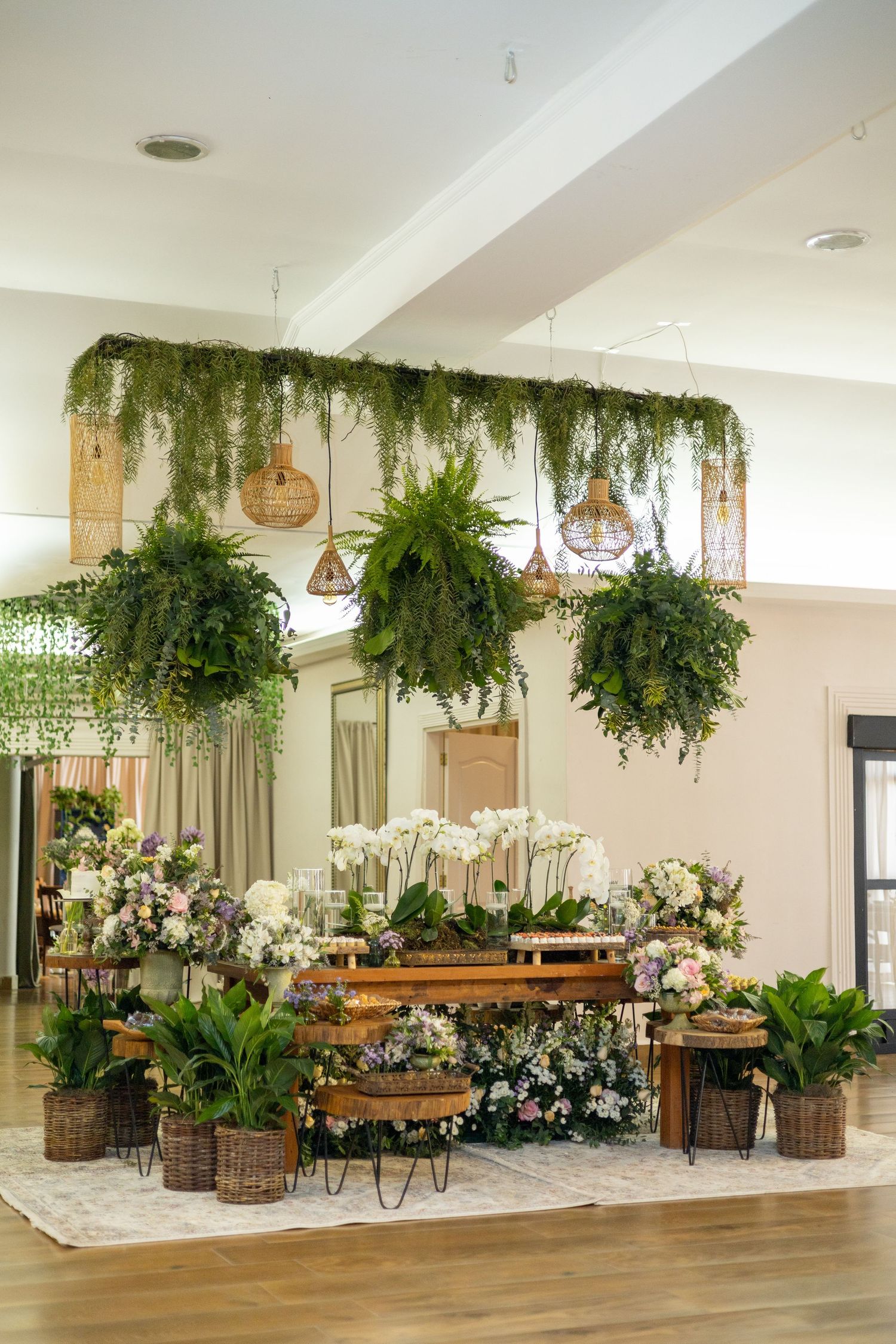 Elegant indoor garden display with hanging greenery, orchids, and floral arrangements on rustic wooden tables with wicker baskets.