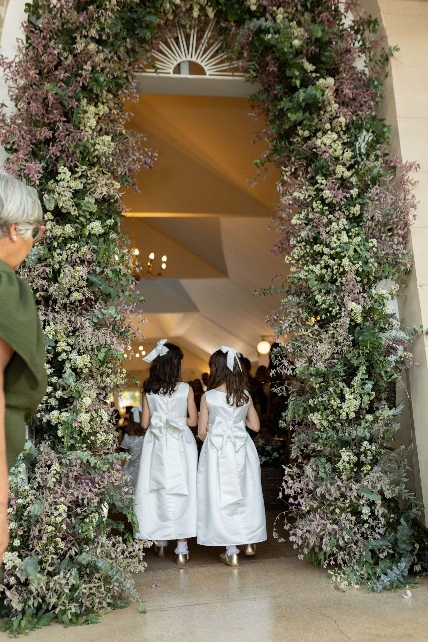 Two flower girls in white dresses walk through an elegant floral arch adorned with greenery and purple blooms.
