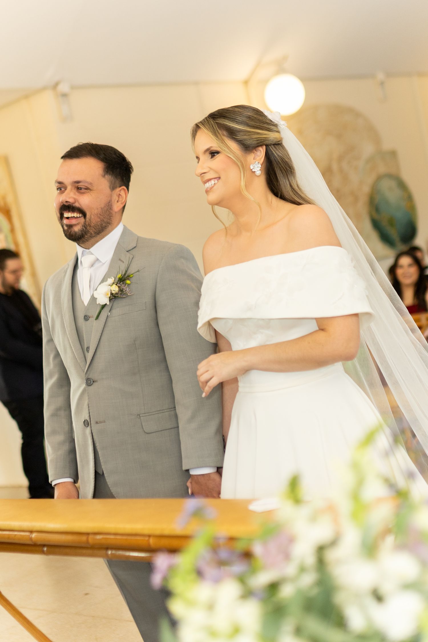 A smiling bride in white off-shoulder gown and veil stands with groom in gray suit at wedding ceremony altar.