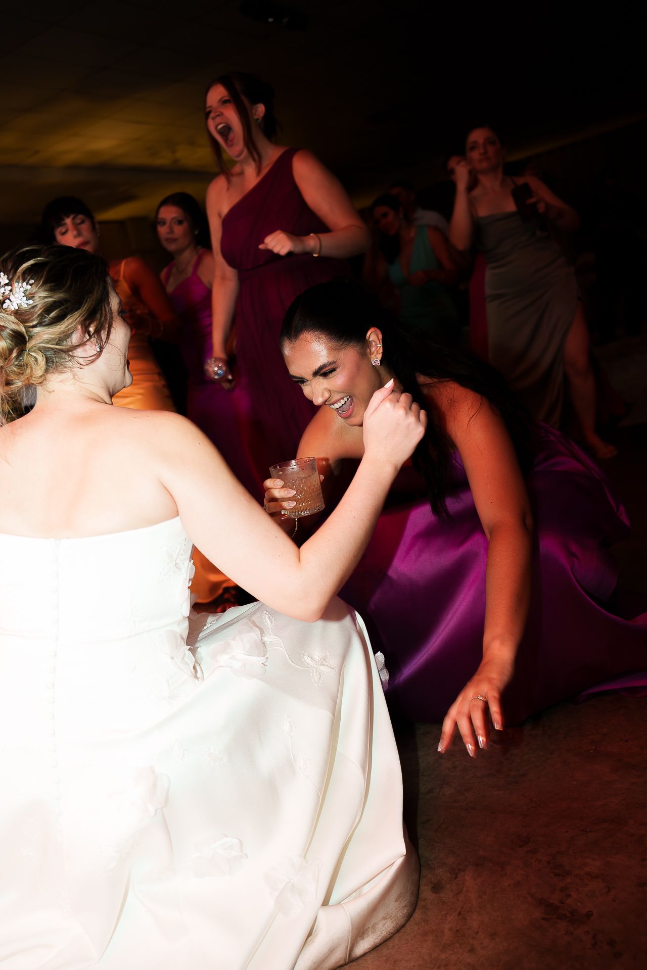 Bride in white gown dancing with bridesmaid in purple dress at lively wedding reception celebration.