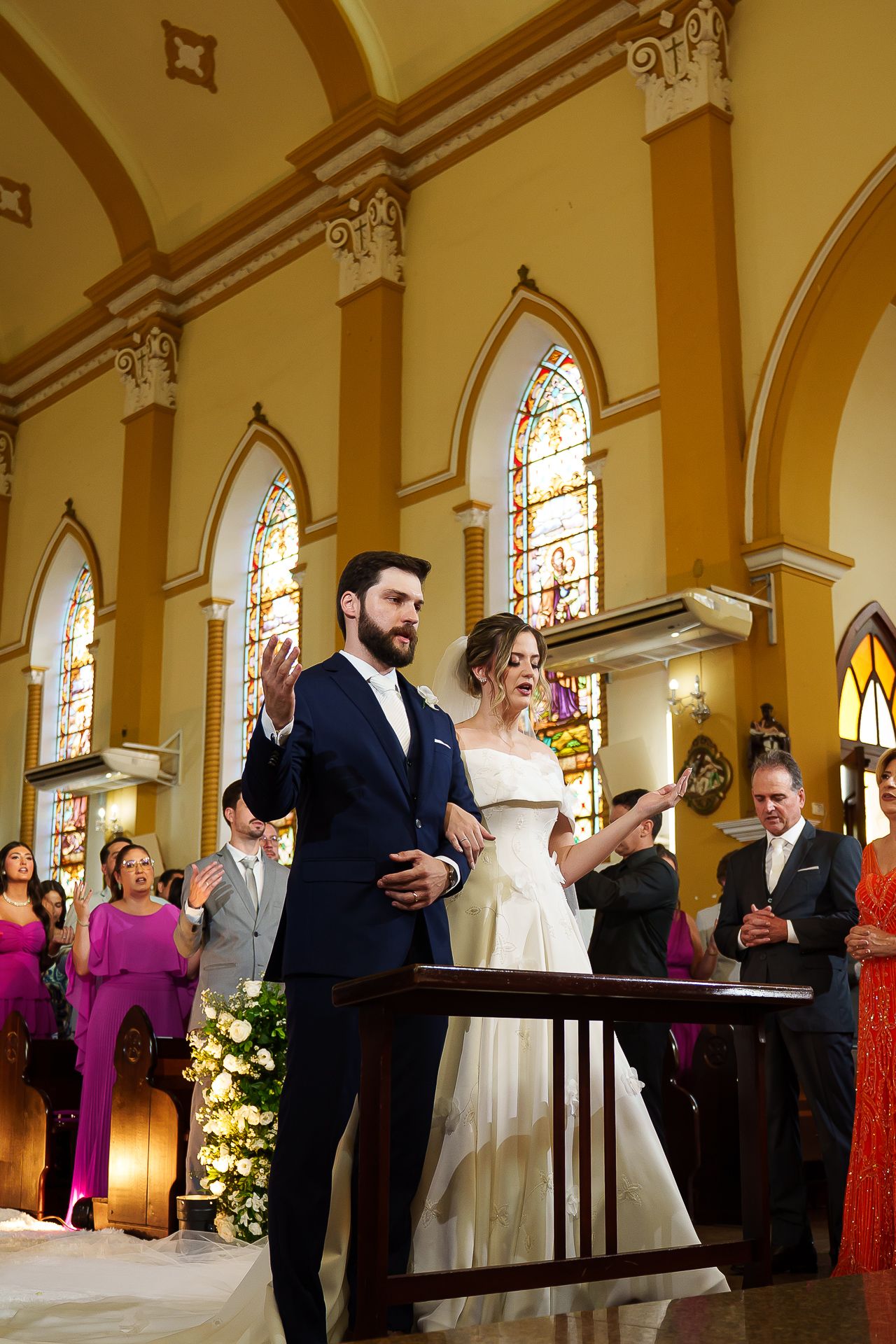 Bride and groom stand at church altar during wedding ceremony, surrounded by guests in elegant attire and stained glass windows.