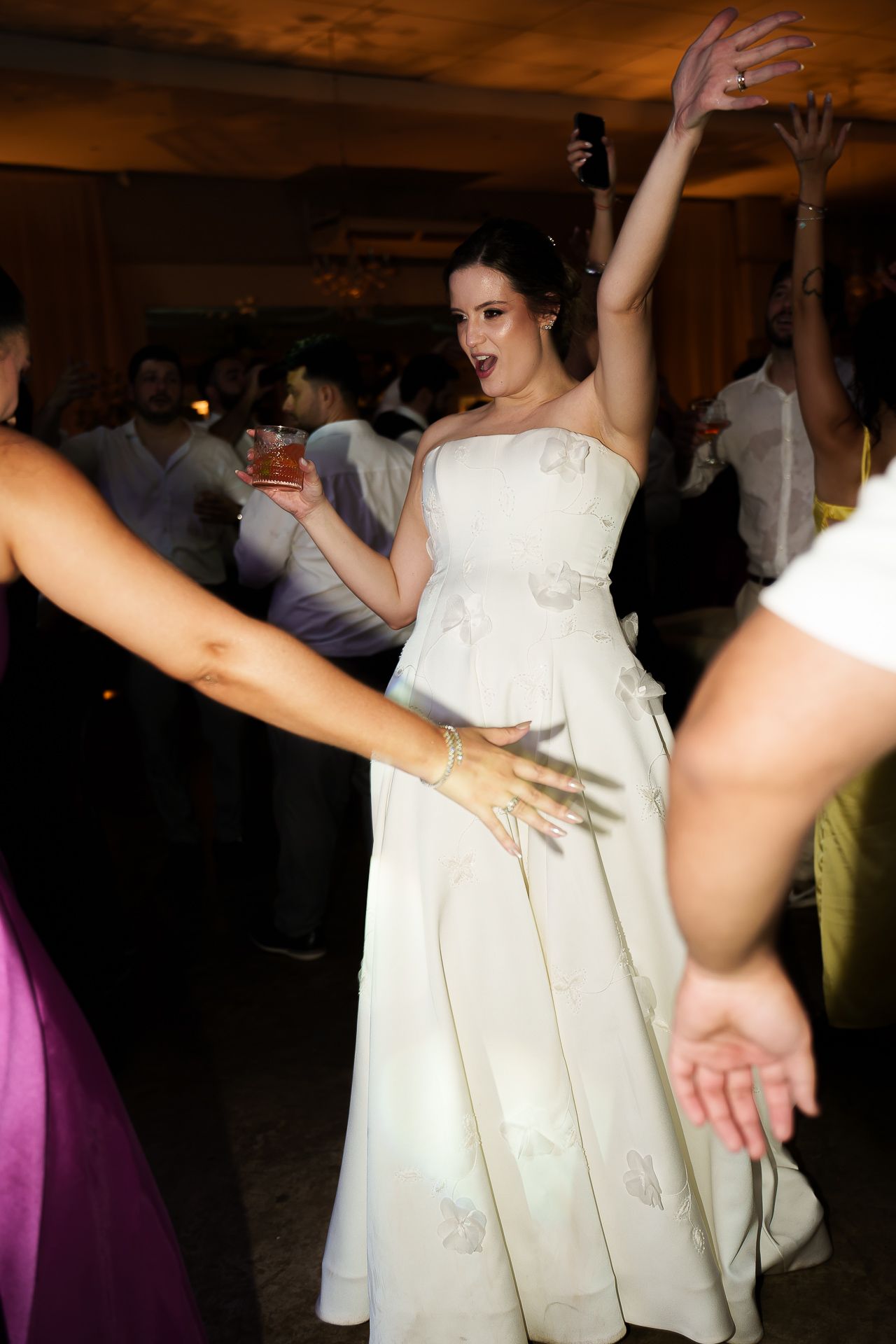 Bride in white gown dancing joyfully at wedding reception, raising arms with drink in hand among guests.