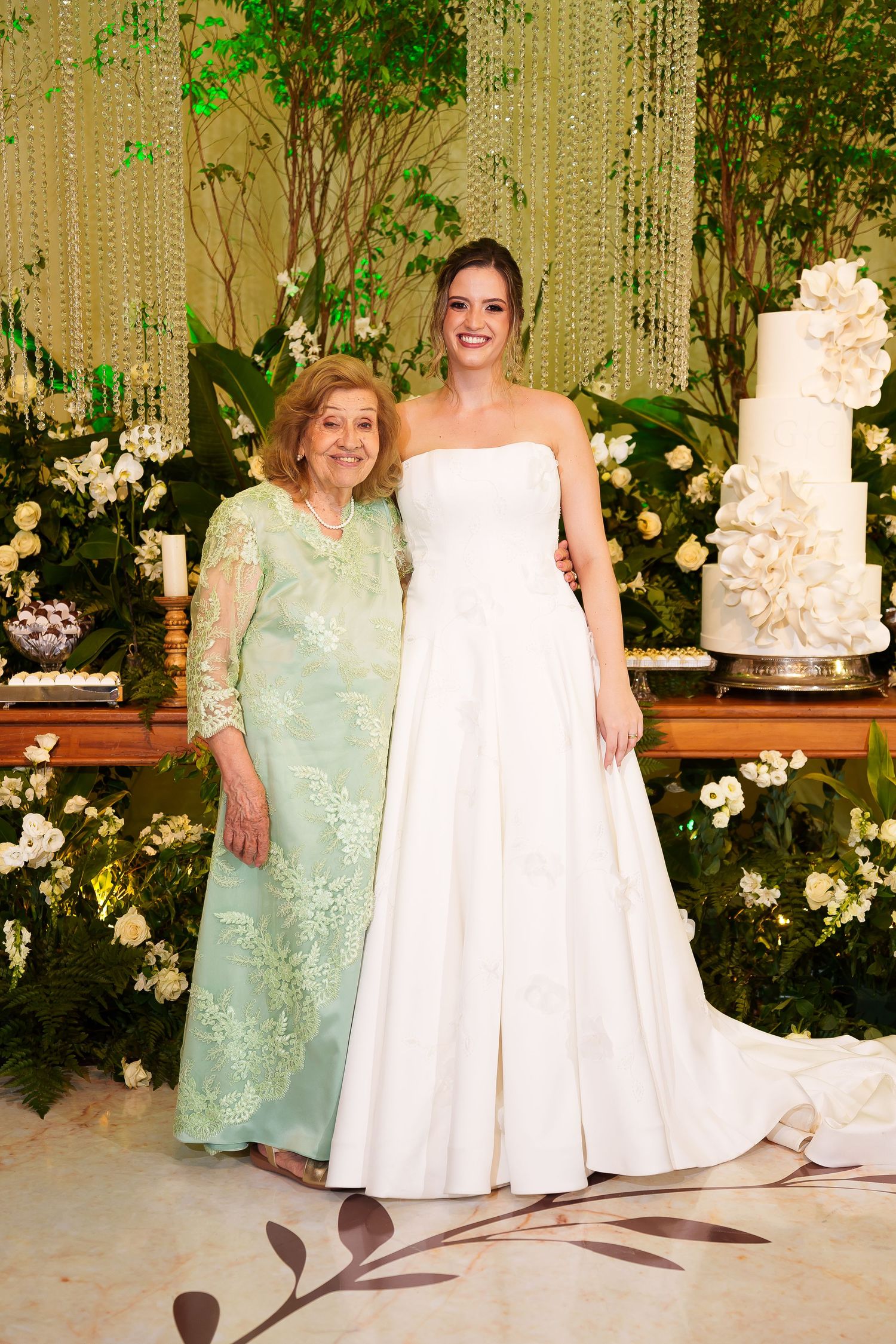 Bride in strapless white gown poses with older woman in green lace dress beside elegant wedding cake.