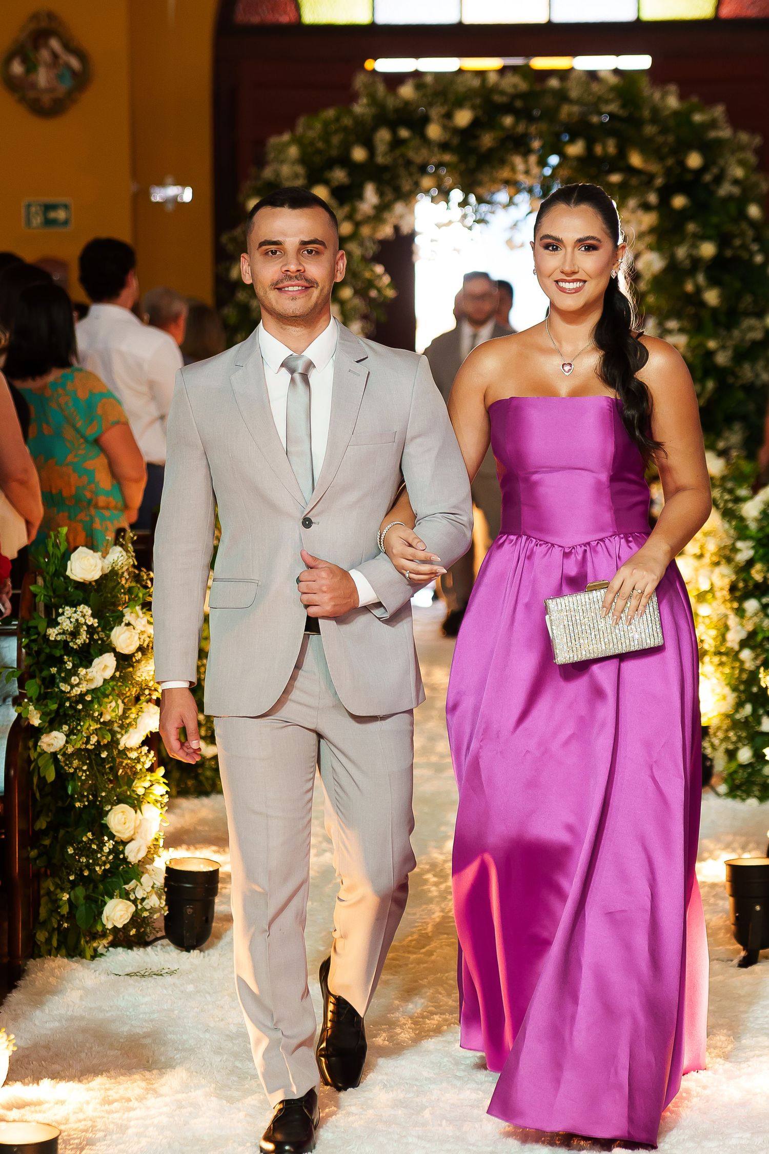 Elegant couple walking down wedding aisle, man in gray suit, woman in strapless purple gown holding silver clutch.
