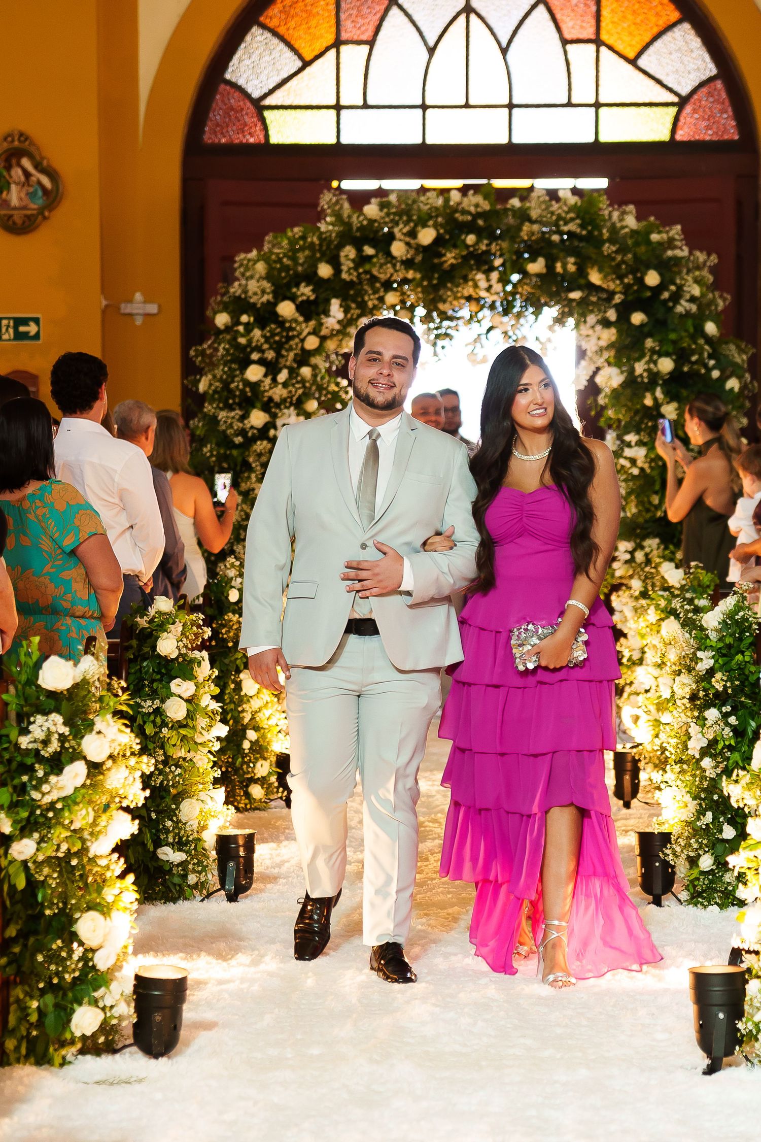 Couple in formal attire walking down floral-decorated wedding aisle, man in light suit, woman in pink tiered gown.