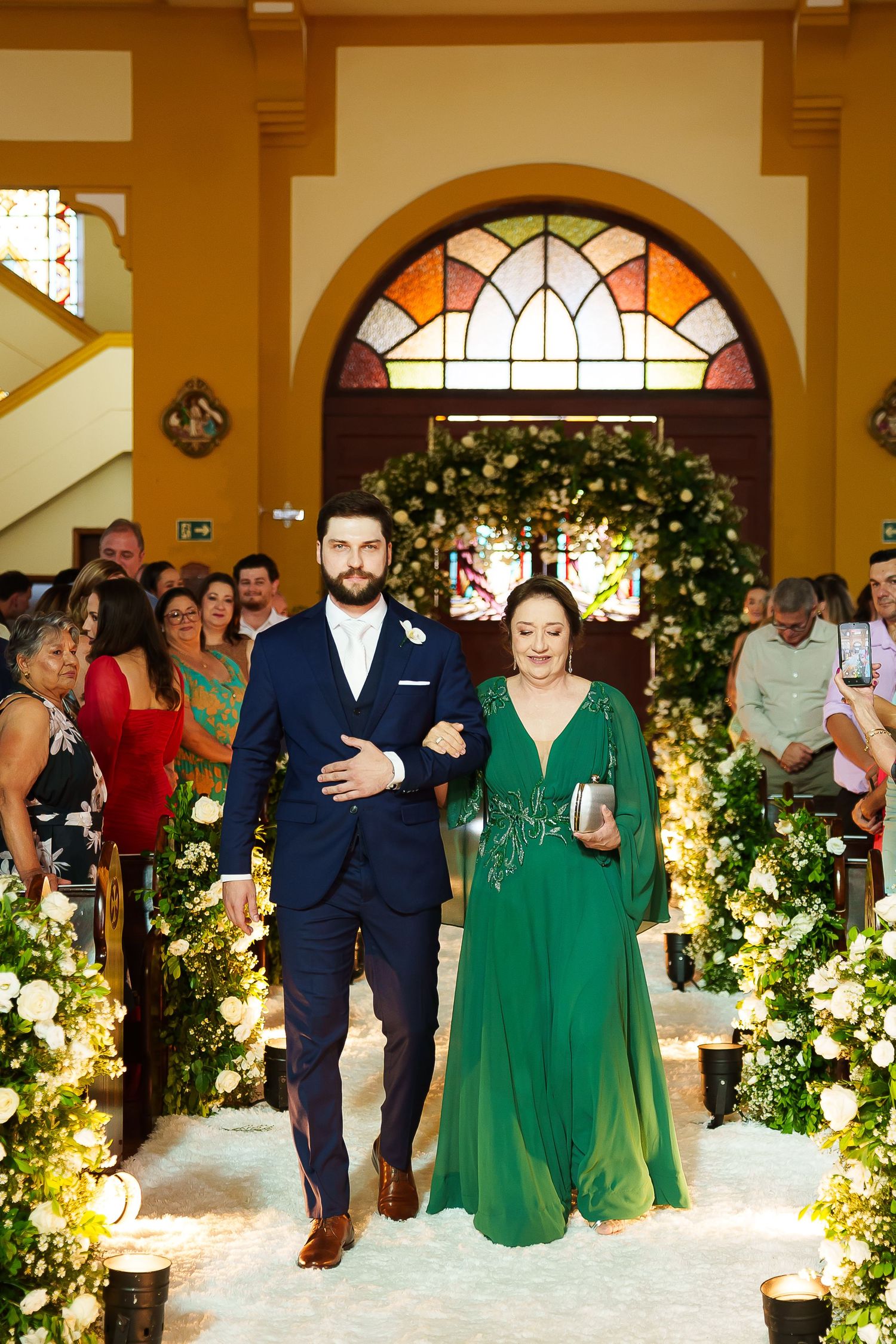 Groom in navy suit walks down church aisle with woman in elegant green gown at wedding ceremony.
