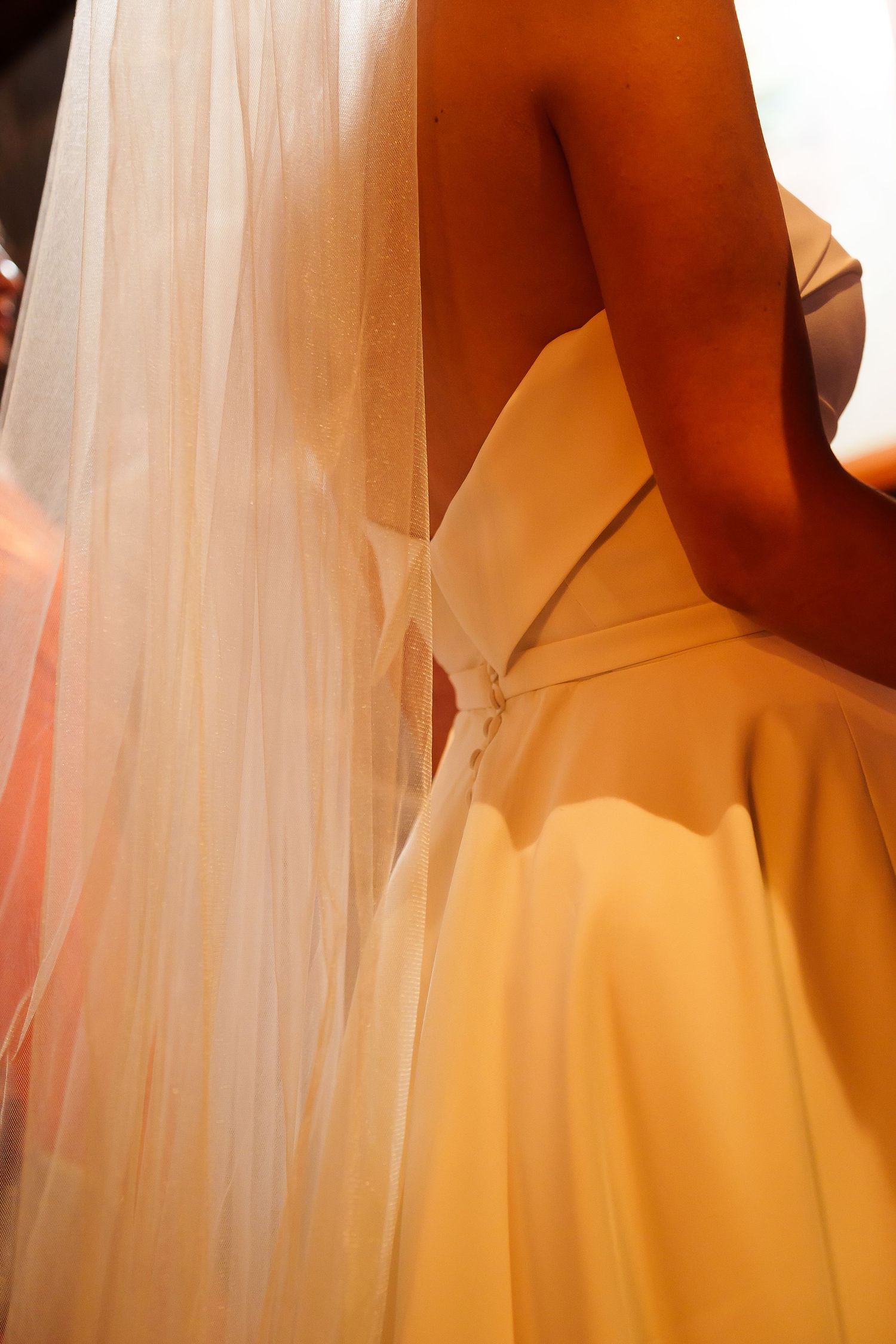 Close-up back view of a bride in a white wedding gown with a flowing veil, illuminated by warm golden light.