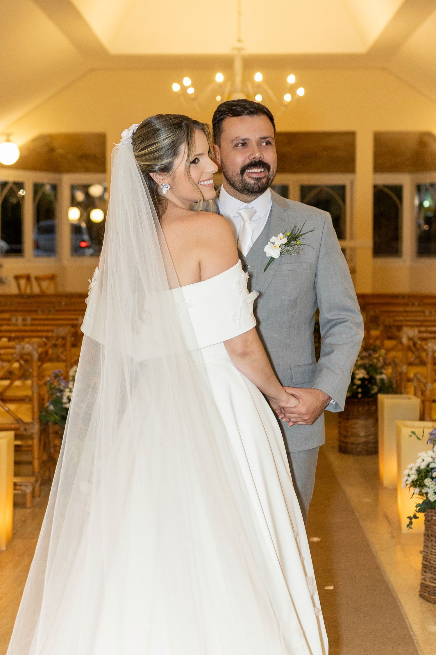 Bride in white off-shoulder gown with veil and groom in gray suit pose together inside an elegant church chapel.