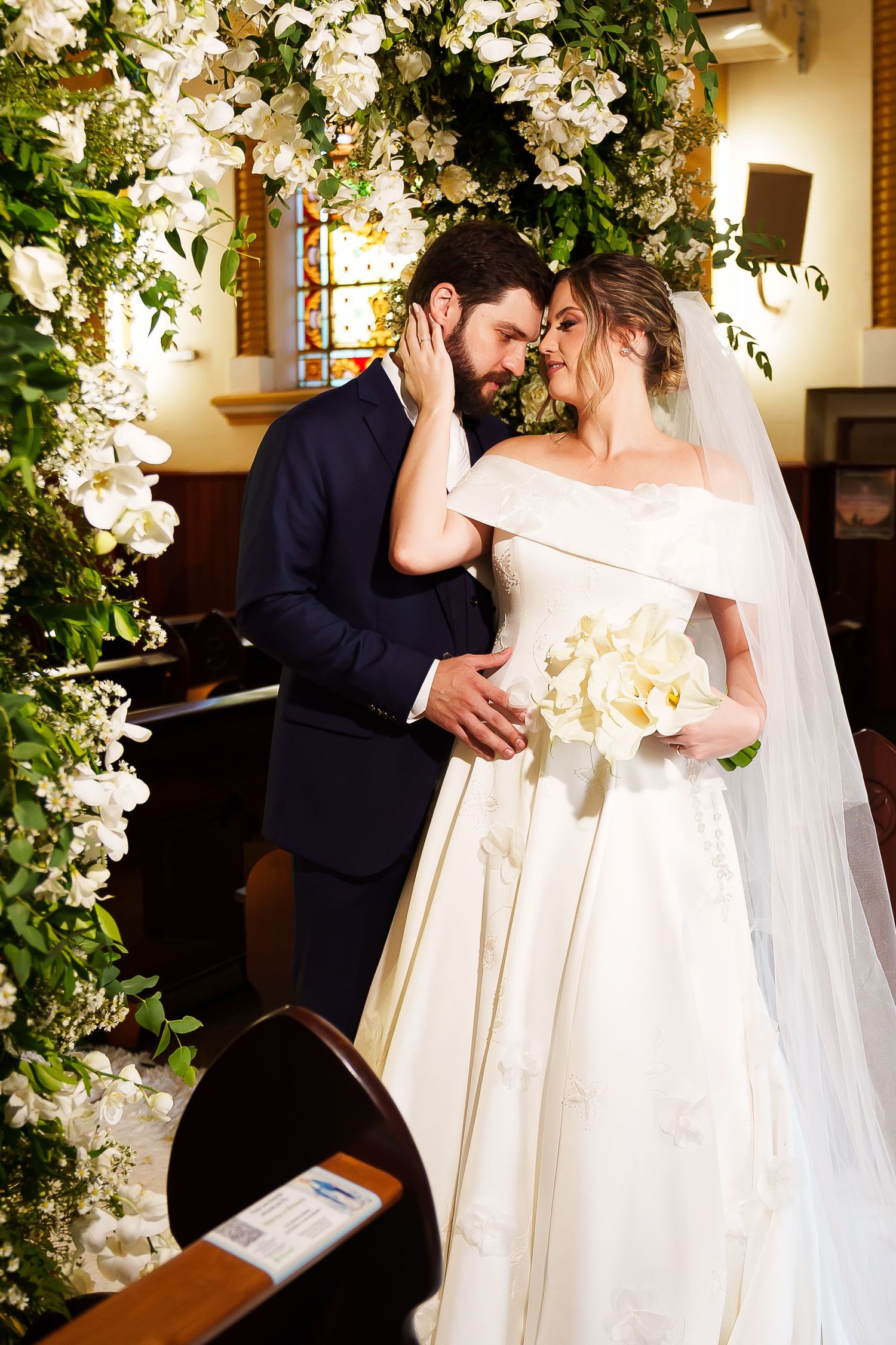 Bride and groom share intimate moment at church altar, surrounded by lush white floral arch, bride holding calla lily bouquet.