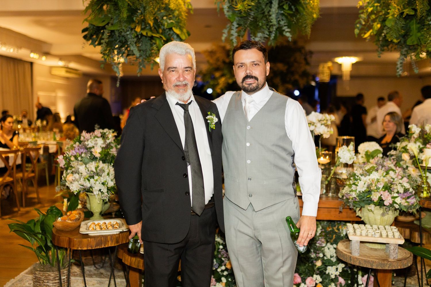 Two men in formal attire pose together at an elegant wedding reception with floral arrangements and greenery.