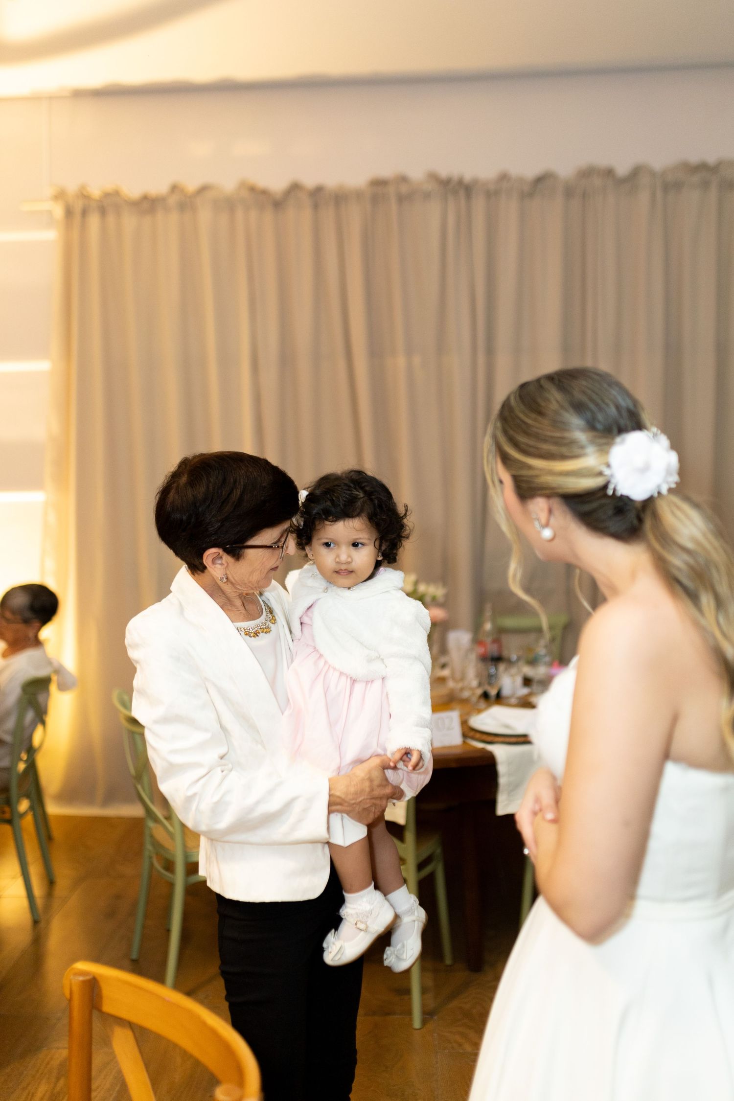 Bride in white dress with flower hair clip talks to older woman holding a baby in pink at a wedding reception.
