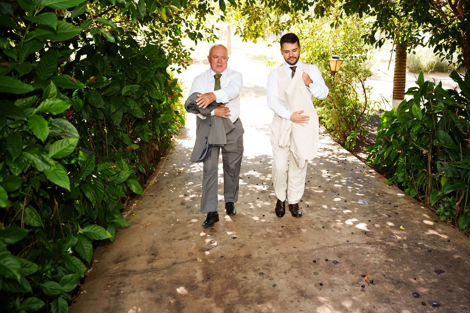 Two men in formal attire walk together down a tree-lined garden path in bright daylight.