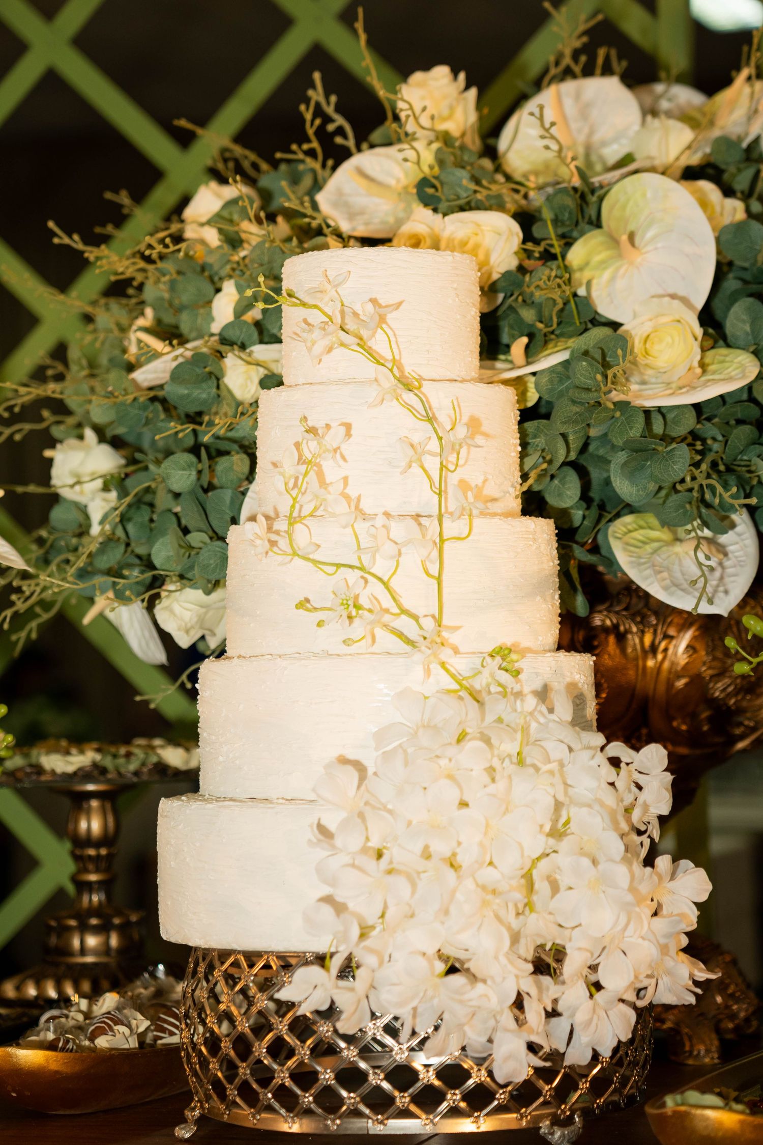 Elegant five-tier white wedding cake adorned with white orchids and gold branches, displayed on a ornate gold stand.