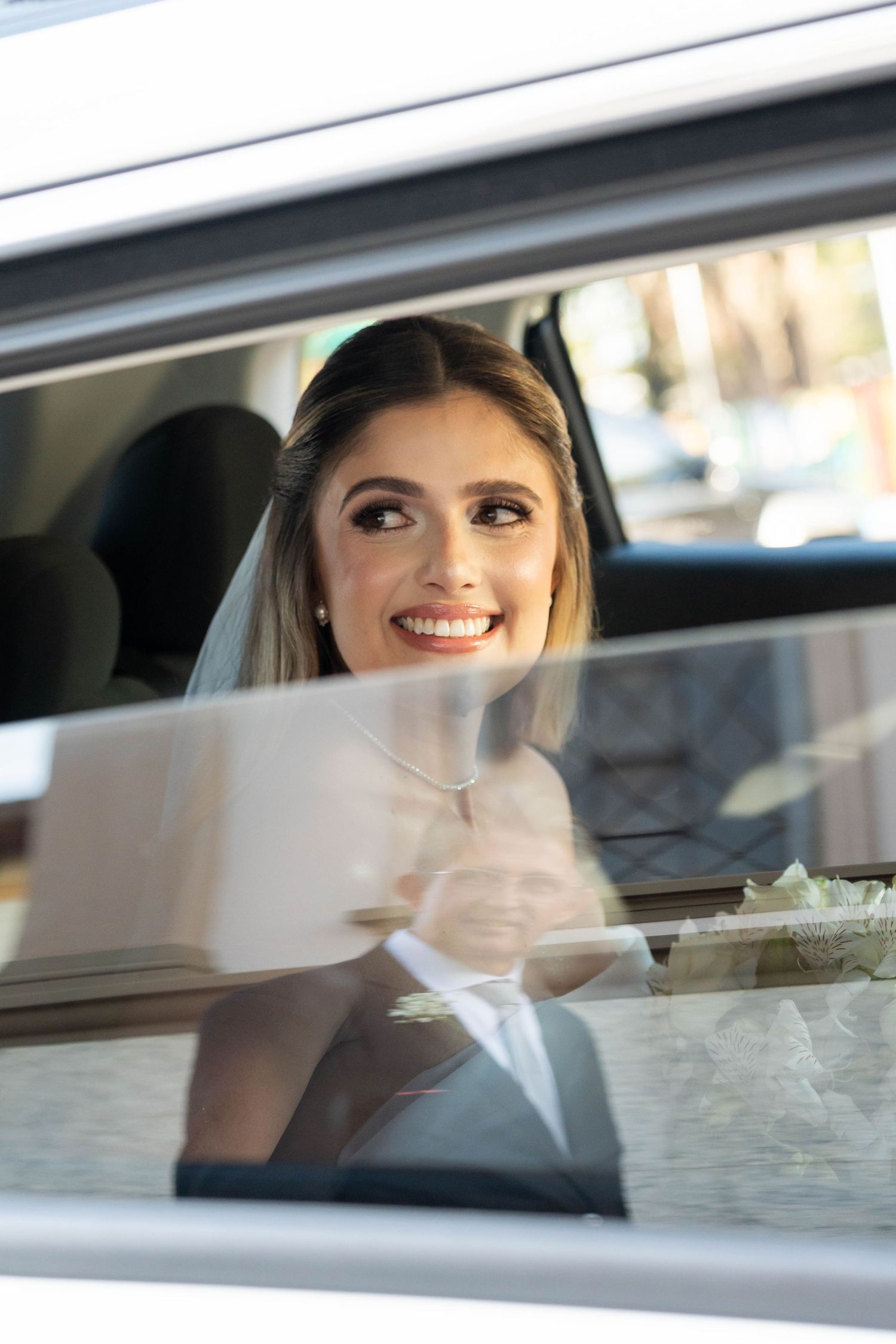 Smiling bride in white veil sitting in a car, holding flowers, with groom reflection visible in window.