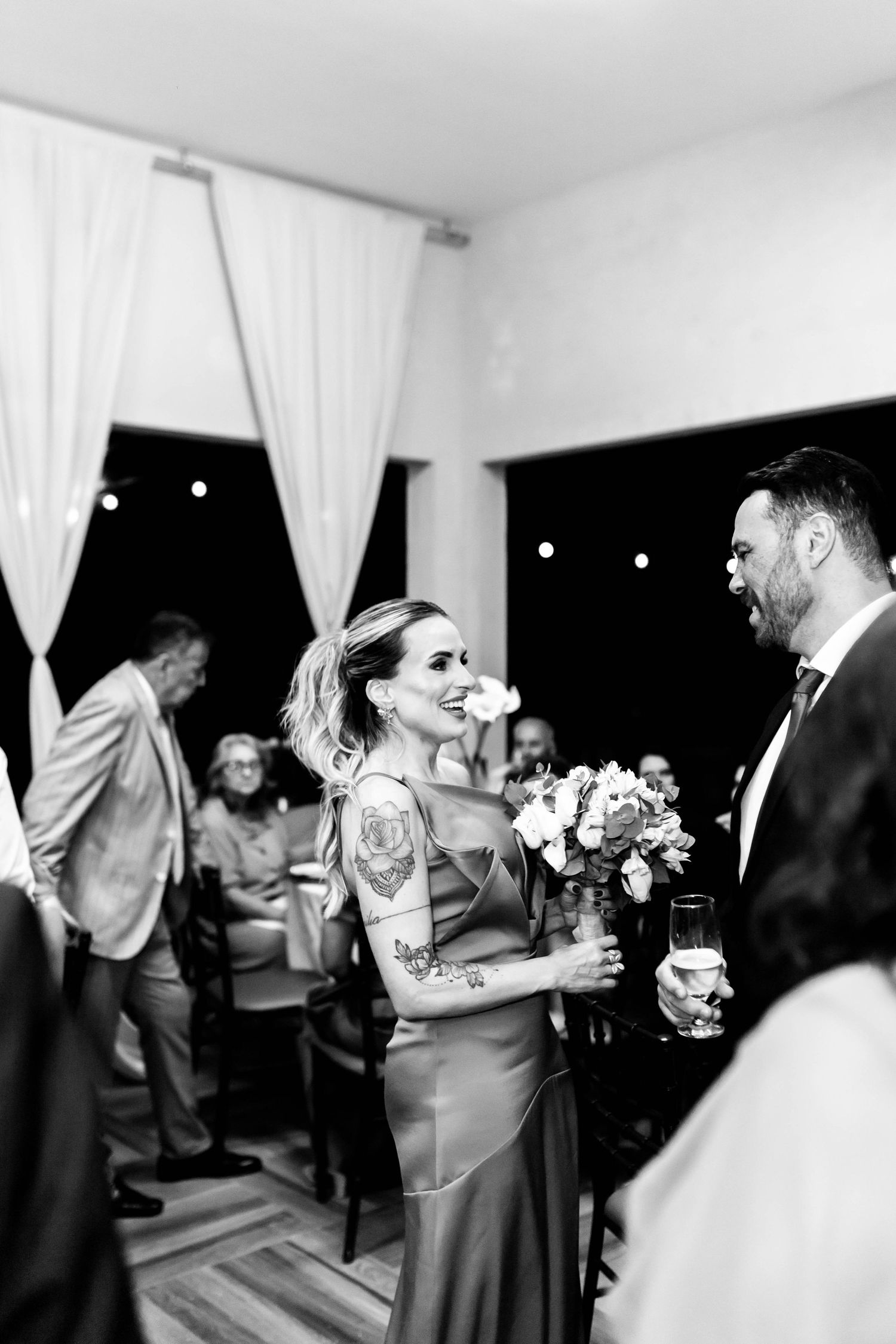 Black and white photo of a tattooed bride in a satin dress smiling at groom during wedding reception.