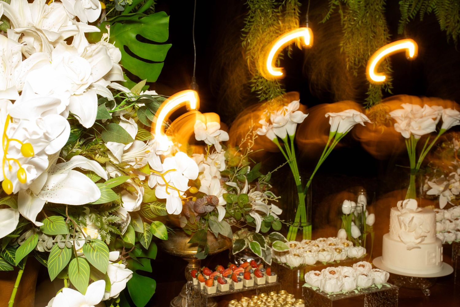 Elegant dessert table with white floral arrangements, glowing neon lights, and assorted sweets at a wedding reception.