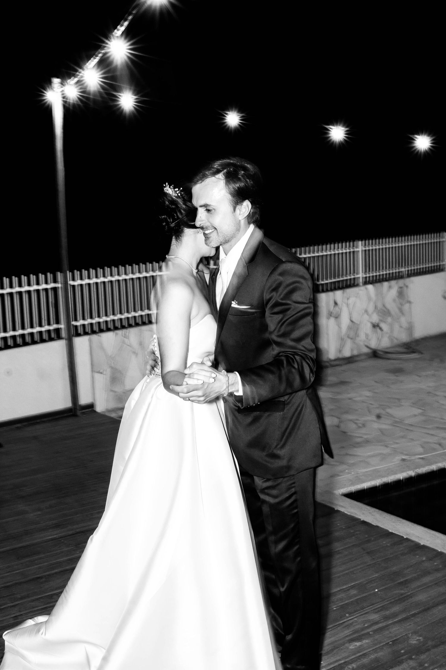 Black and white photo of a bride and groom sharing their first dance outdoors at night under string lights.