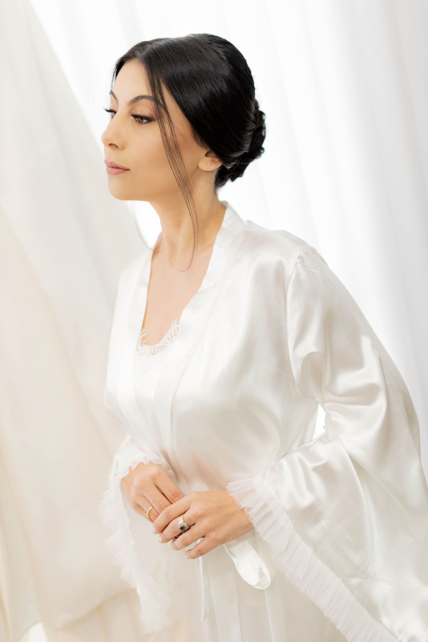 Elegant bride in white satin robe with lace trim, dark hair updo, standing gracefully near sheer white curtains.