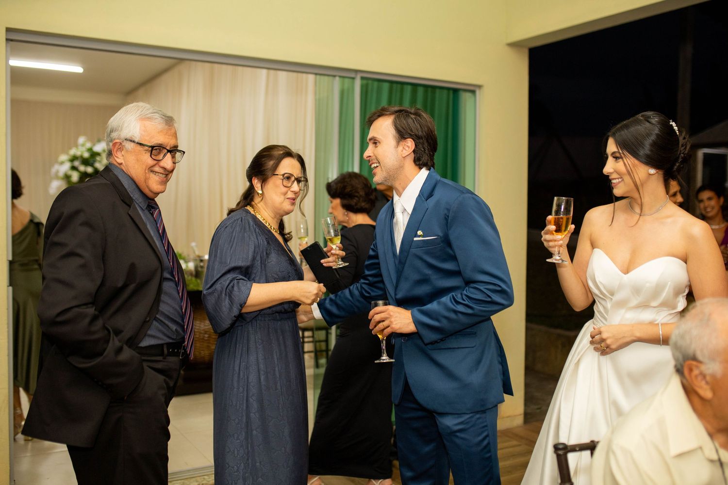 Bride in white gown and groom in blue suit toasting champagne with guests at elegant wedding reception.
