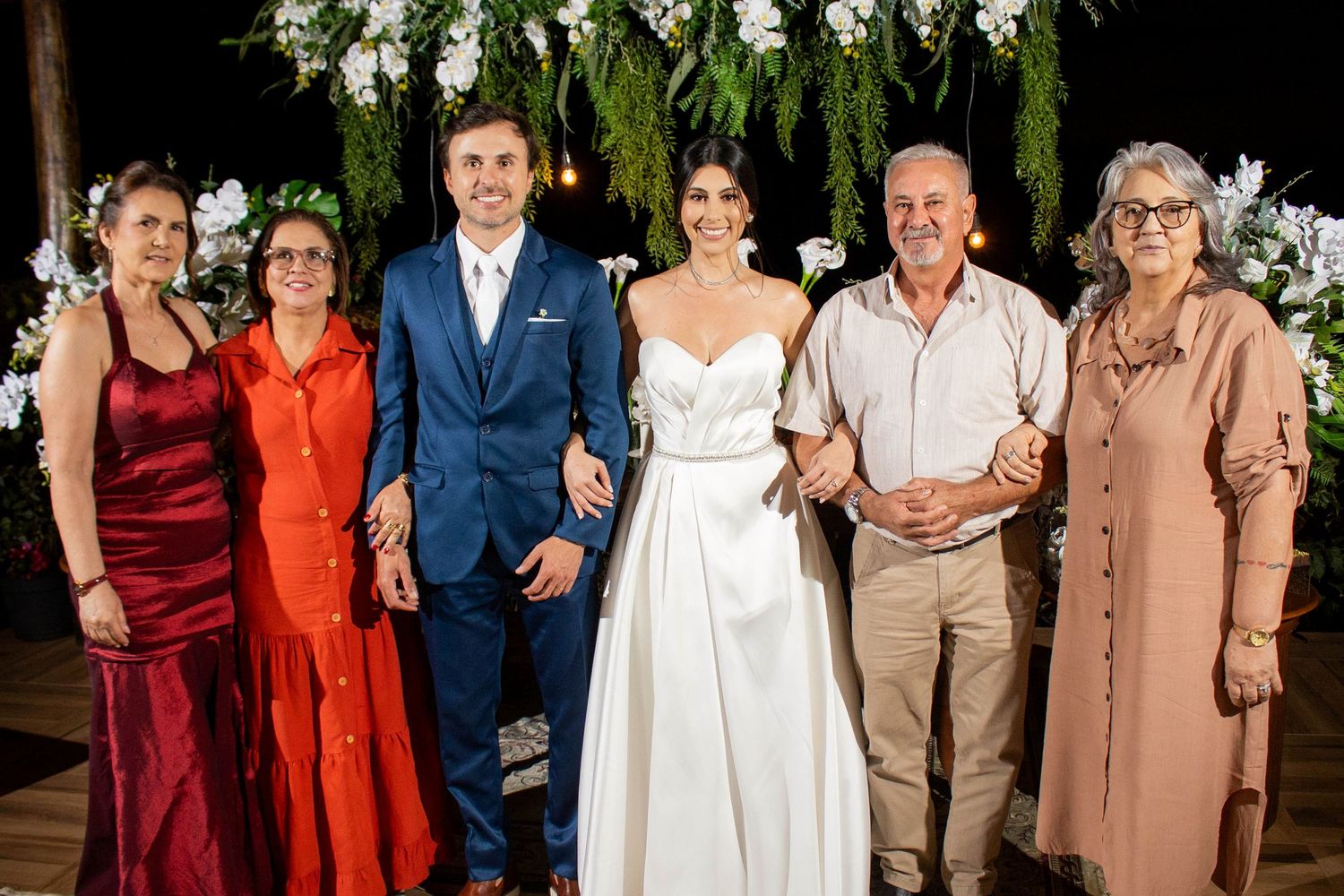Wedding couple in white and blue attire poses with four family members under floral arch at evening reception.