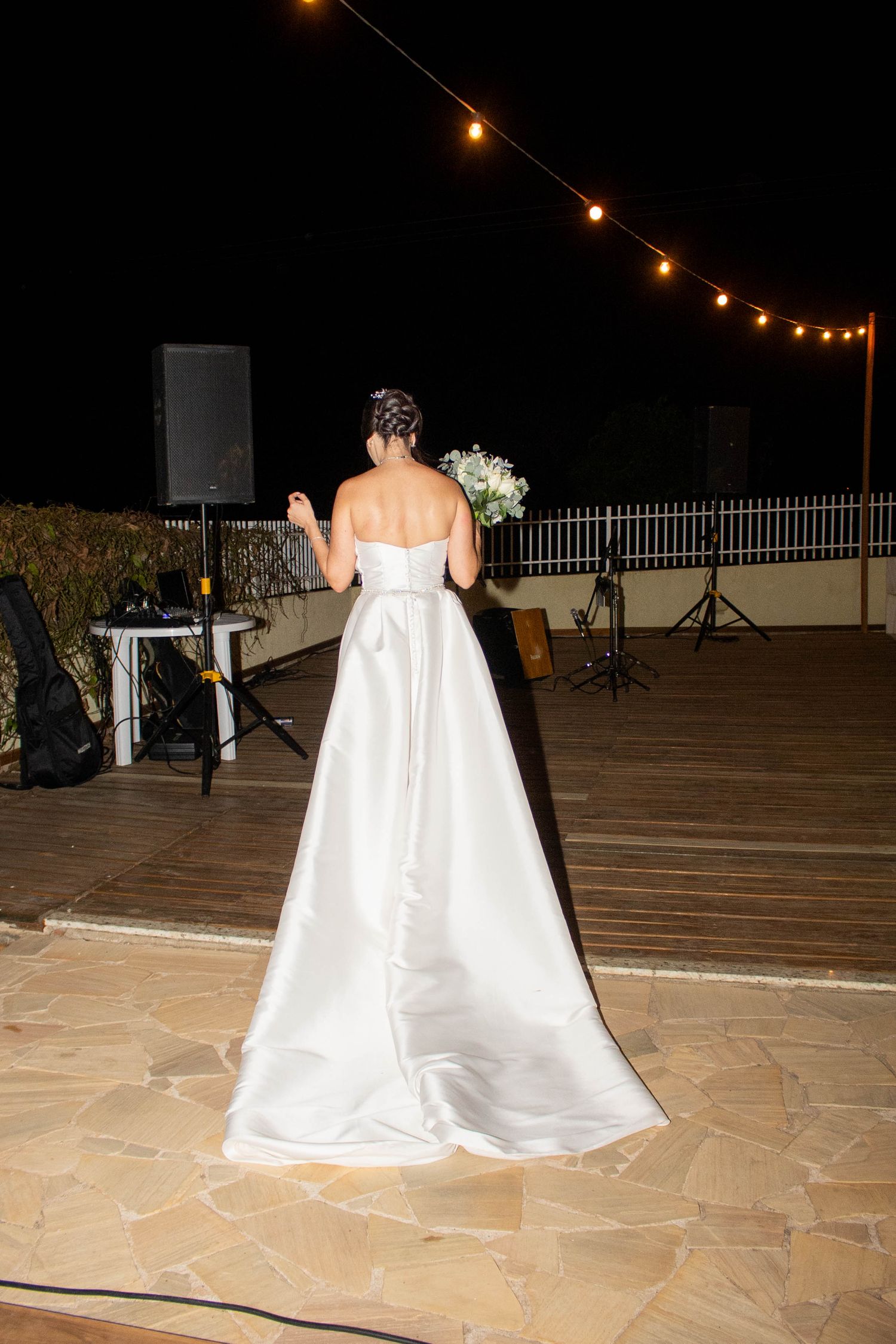 Bride in white strapless gown holds bouquet on outdoor dance floor at night, string lights glowing above.