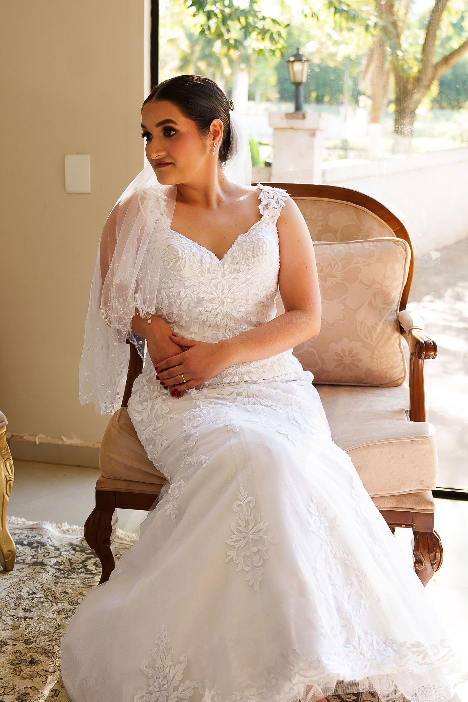 Bride in white lace mermaid gown and veil sits gracefully on ornate gold chair, gazing thoughtfully to the side.