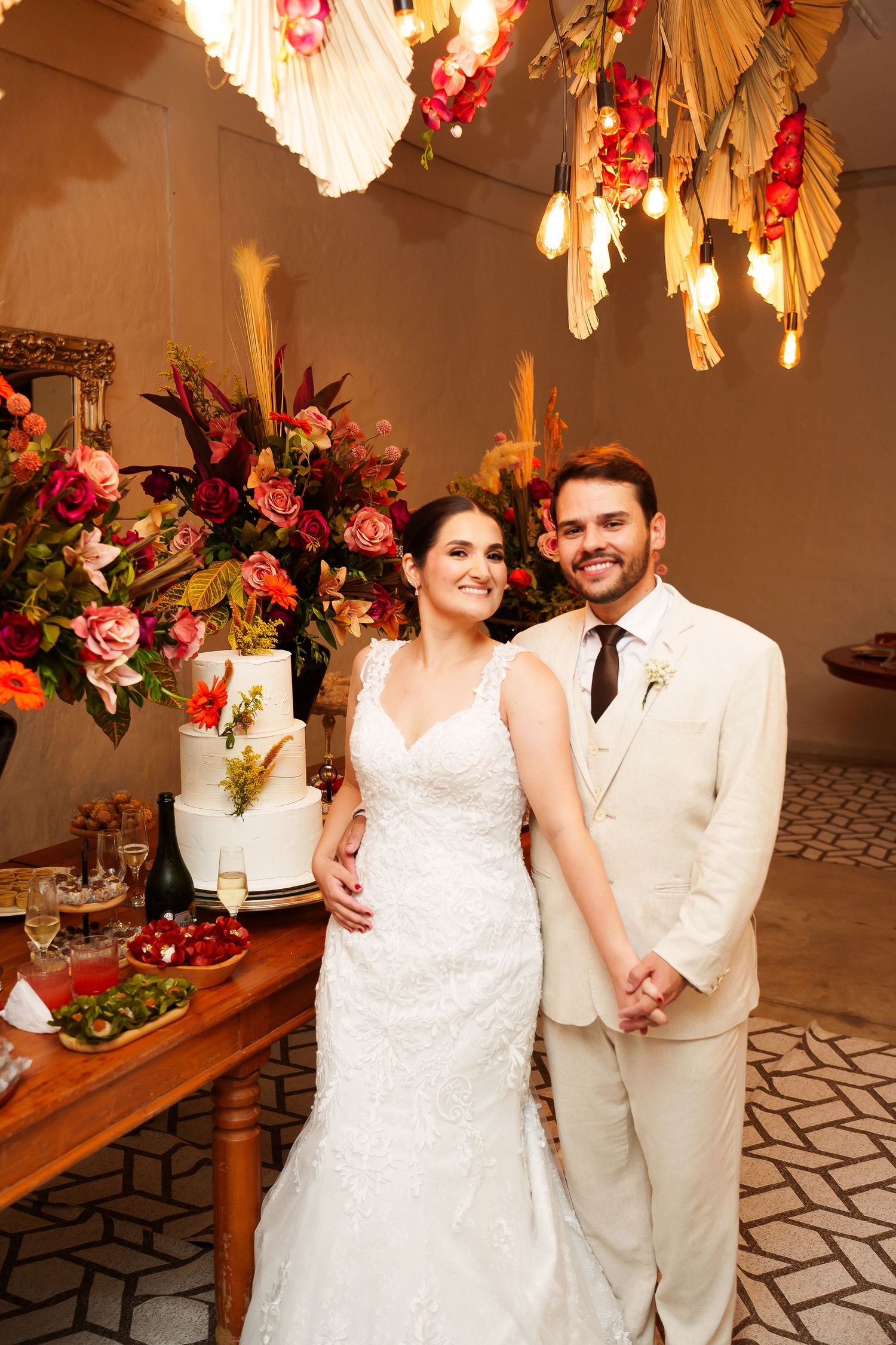 Smiling bride and groom pose together at their wedding reception beside a floral arrangement and white tiered cake.