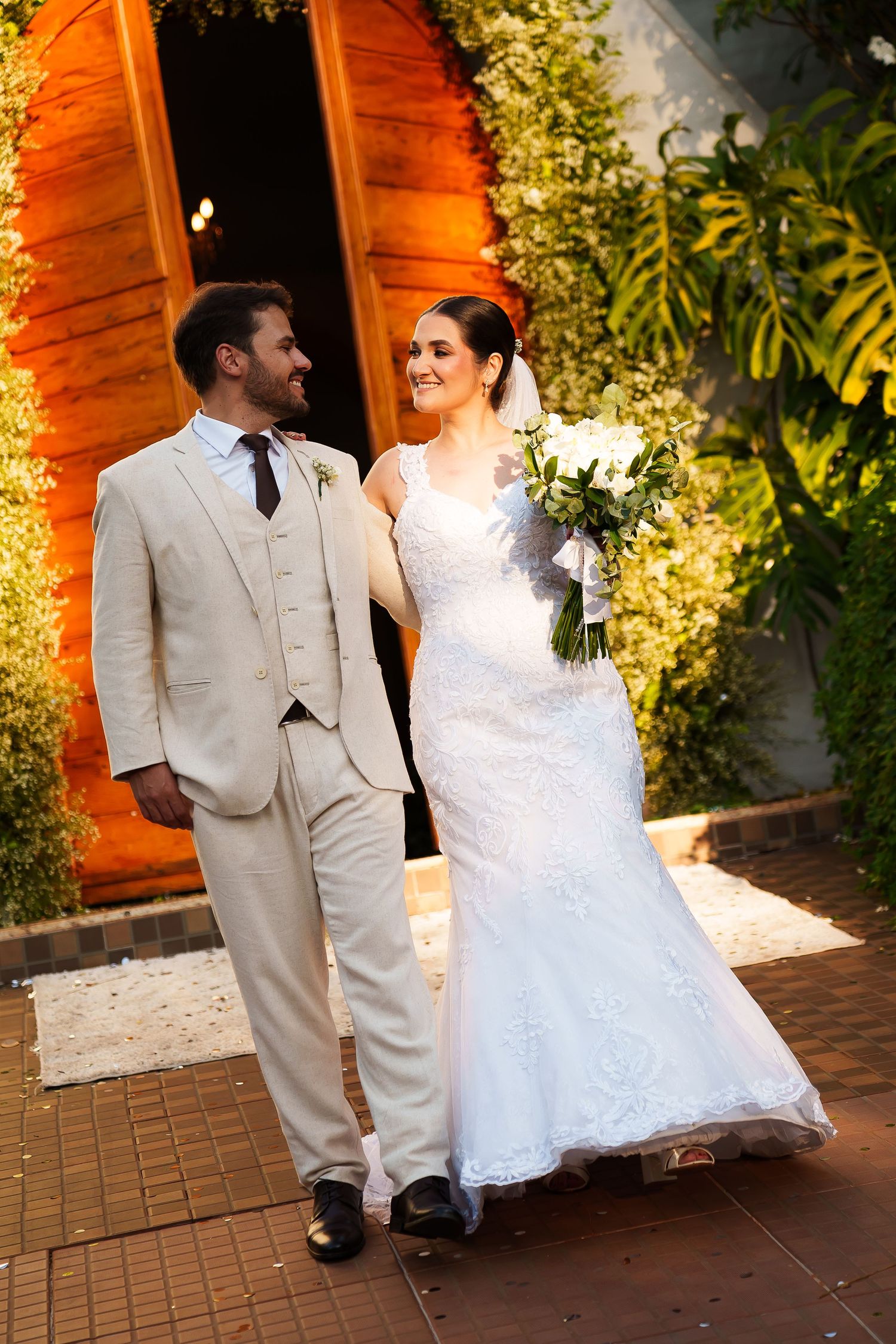 Bride in white lace gown holding bouquet walks with groom in beige suit at outdoor wedding venue.