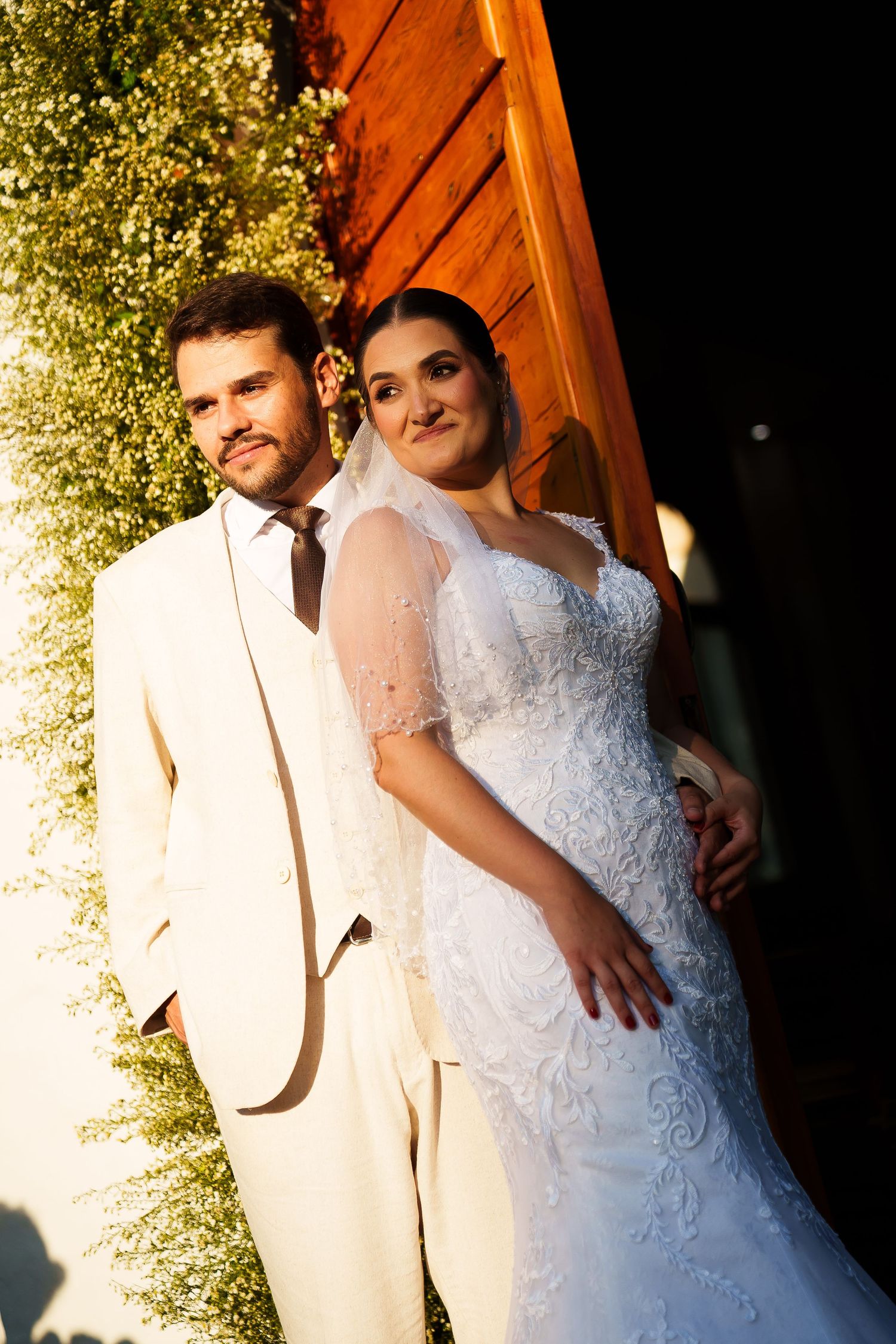 Elegant bride in lace wedding gown and groom in cream suit posing together outdoors in golden sunlight.