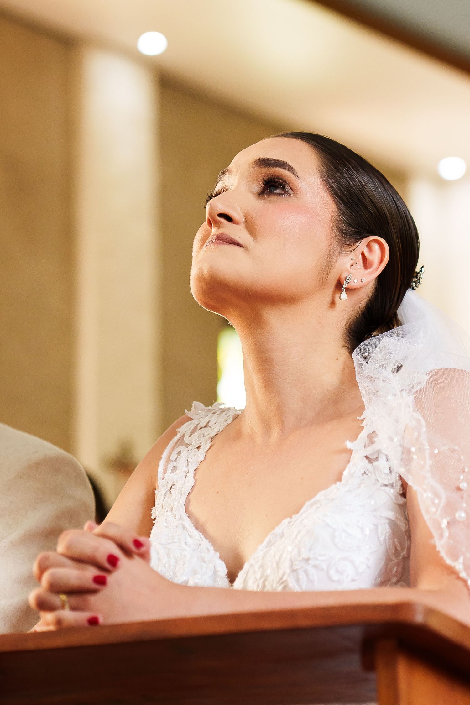 Bride in white lace gown praying with hands clasped at church pew, looking upward with veil and red nails.
