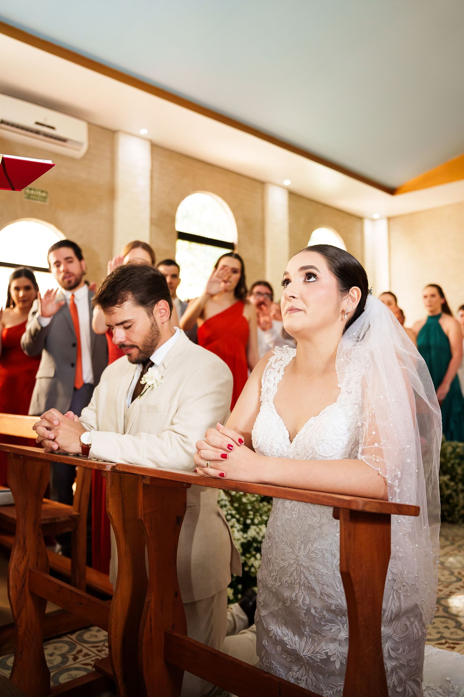 Bride and groom kneeling in prayer at church altar during wedding ceremony, guests waving in background.