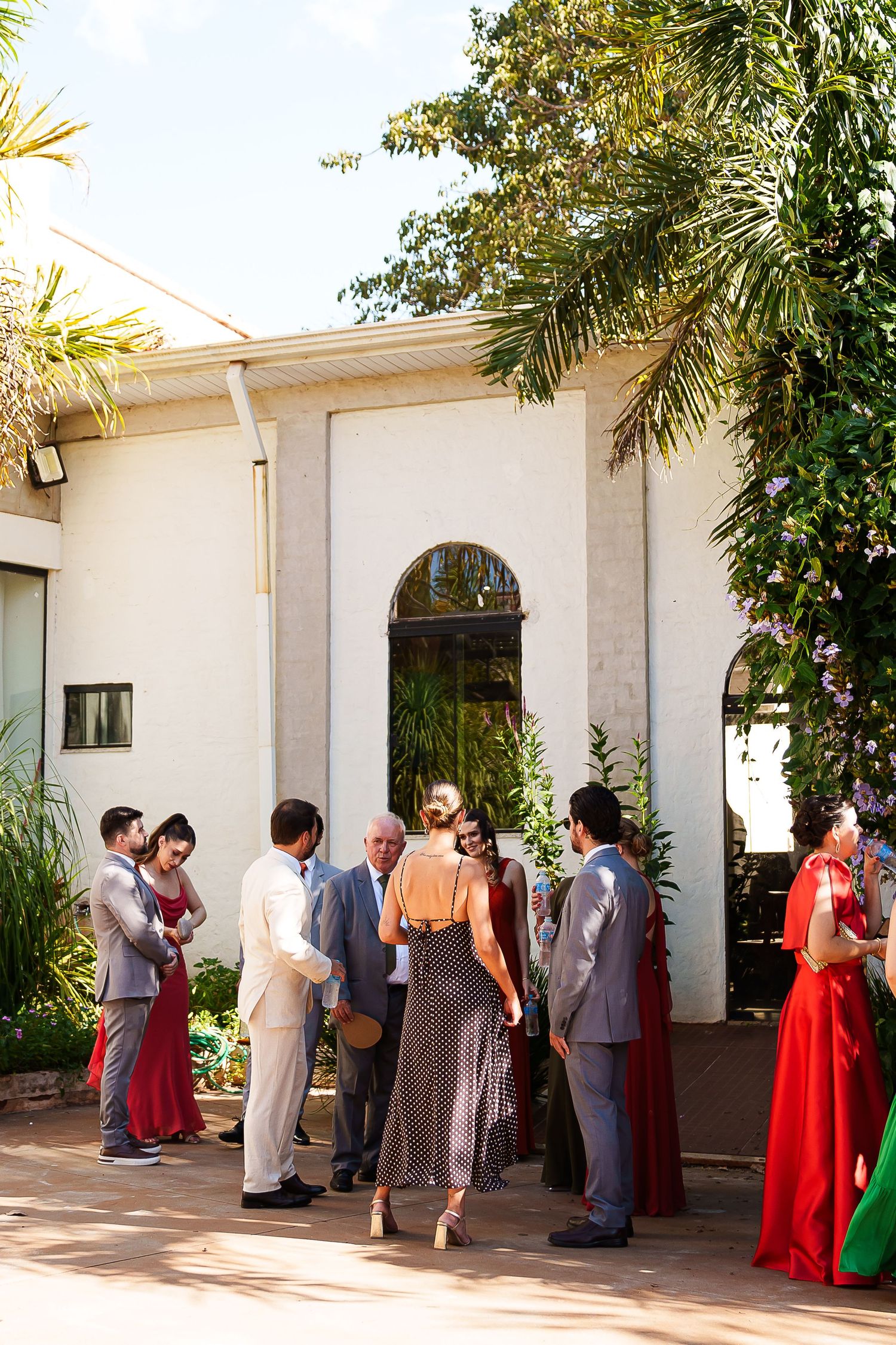 Elegantly dressed wedding guests mingle outside a white venue surrounded by lush tropical trees on a sunny day.