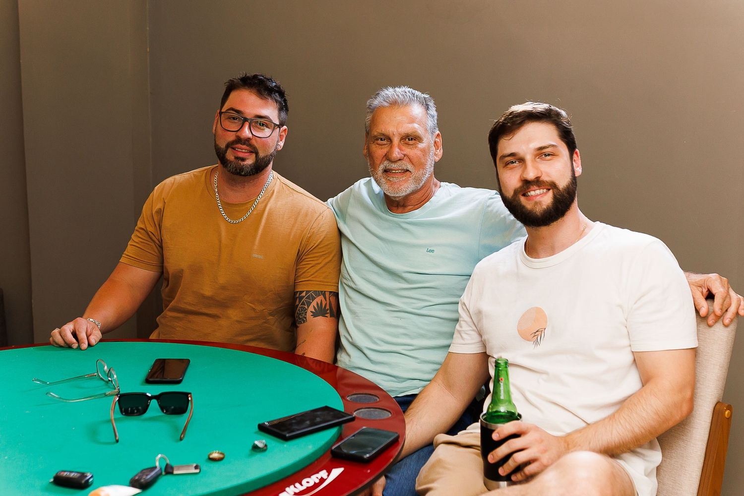Three smiling men pose together at a green poker table, one holding a beer bottle in a casual indoor setting.
