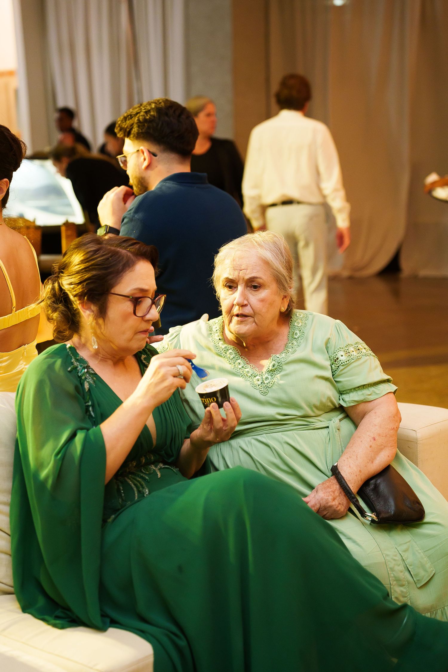 Two women in green dresses sit together at an elegant event, one showing something on her phone to the elderly woman beside her.