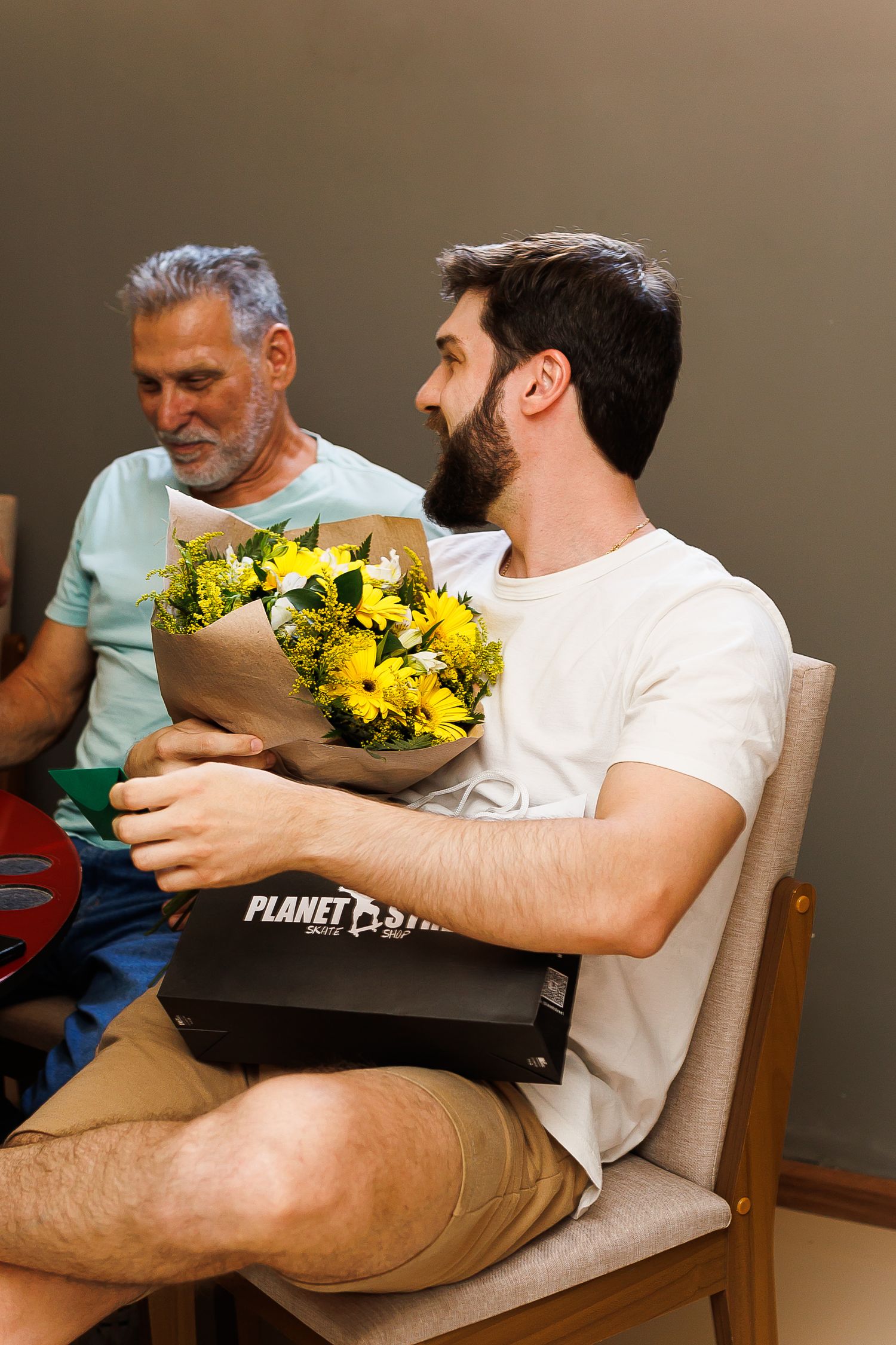 Young bearded man holding yellow flowers bouquet and Planet Fitness box while seated, older man visible in background.