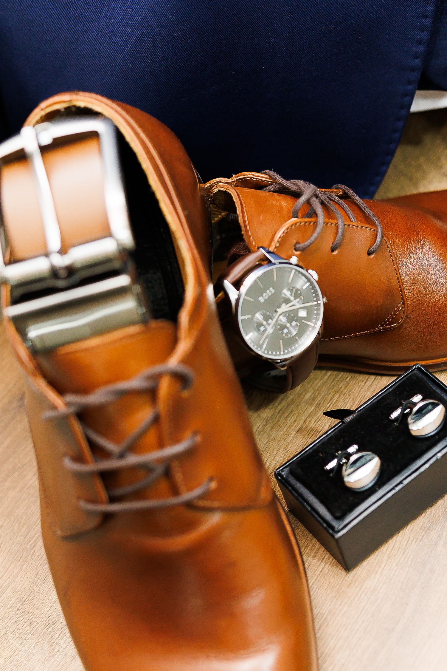 Brown leather dress shoes with matching belt, silver watch, and cufflinks arranged on wooden surface.