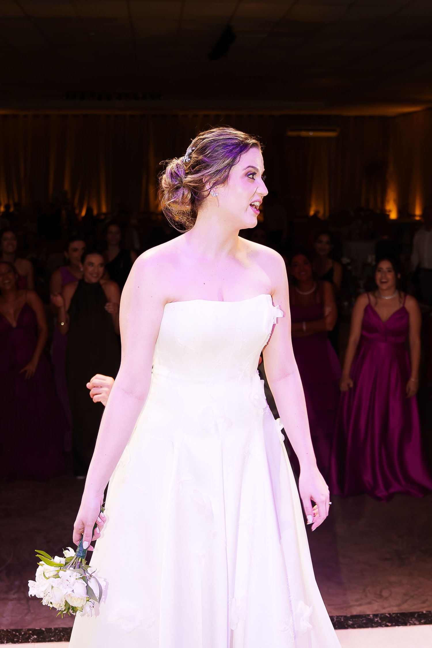 Bride in strapless white gown holds bouquet before tossing it to guests in purple dresses at wedding reception.